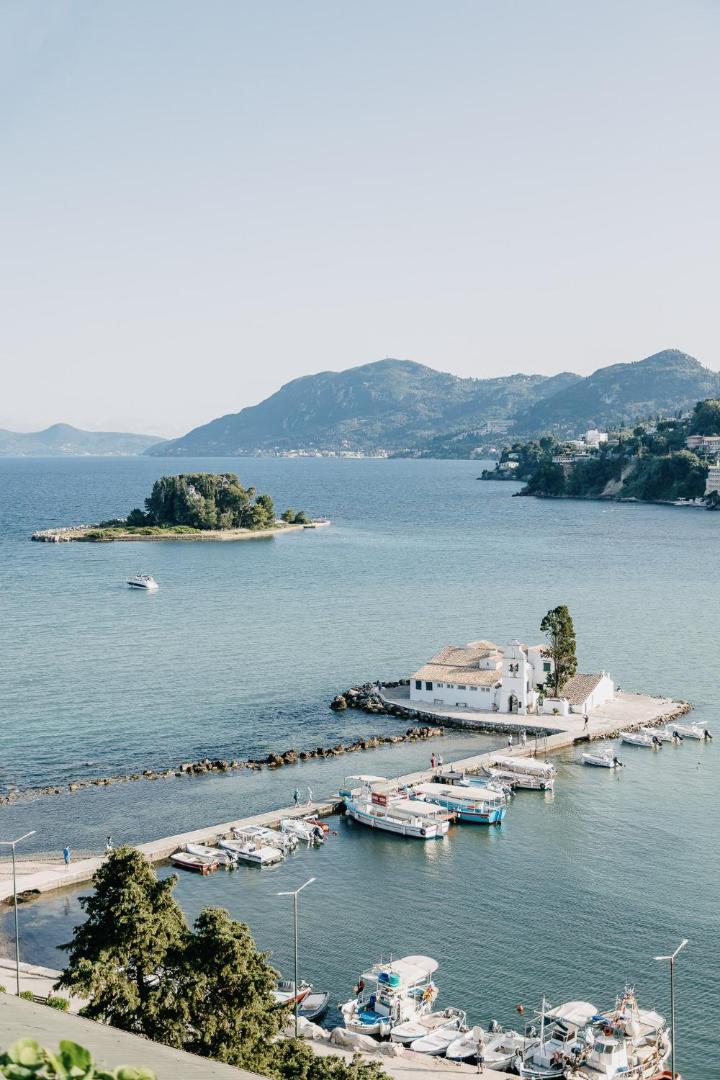 Skats uz Vlacherna klosteri un Pontikonisi salu no Corfu Holiday Palace viesnīcas Korfu salā — zila jūra un kalni fonā