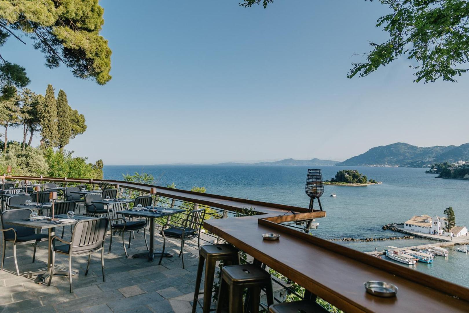 Viesnīcas Corfu Holiday Palace restorāna terase ar galdiņiem un skatu uz Pontikonisi salu un jūras līci
