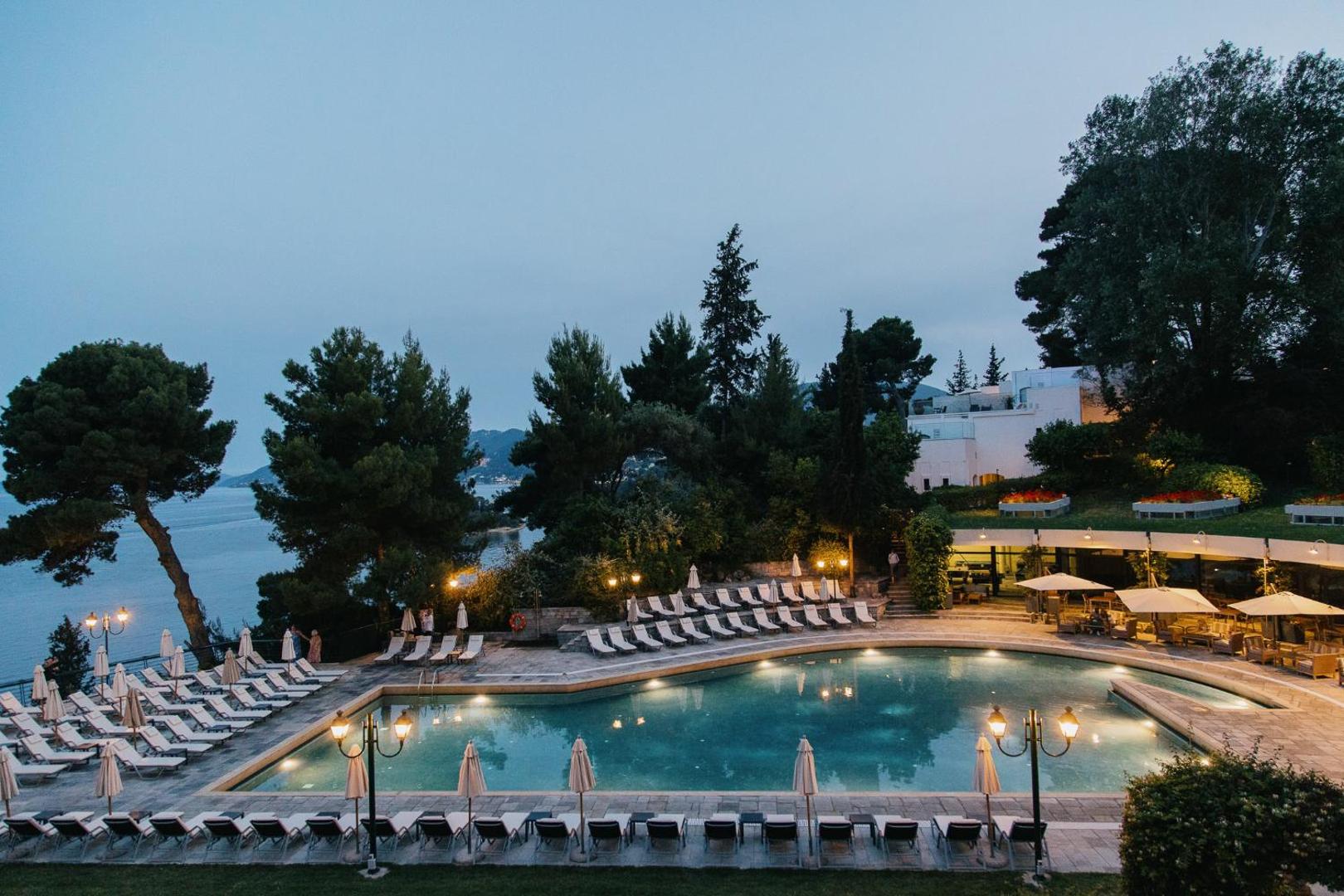 Corfu Holiday Palace baseina zona vakarā ar iedegtām gaismām, atpūtas krēsliem un skatu uz jūru krēslas stundā Kanoni reģionā