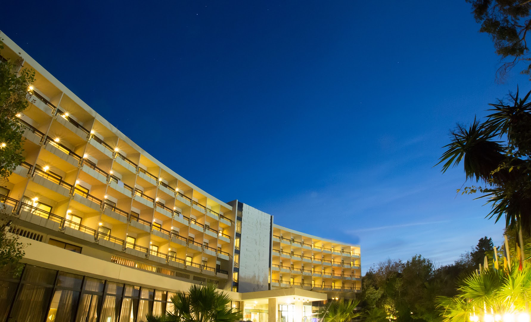 Corfu Holiday Palace viesnīcas modernā arhitektūra un izgaismotie numuru balkoni pret zilo vakara debesu fonu Korfu.