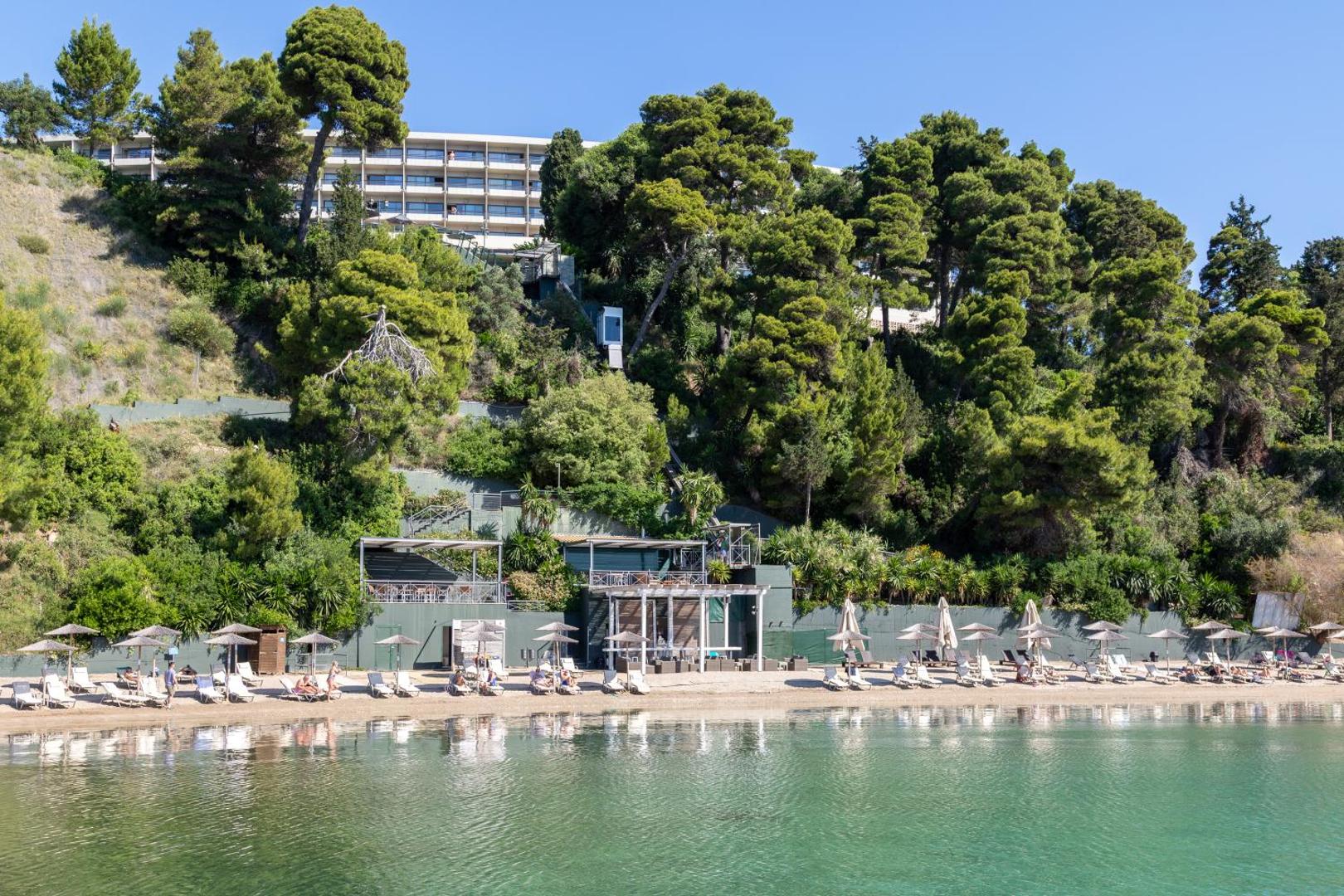 Corfu Holiday Palace privātā smilšu pludmale ar saulessargiem un zviļņiem pie zaļojoša kalna Korfu, Grieķijā.