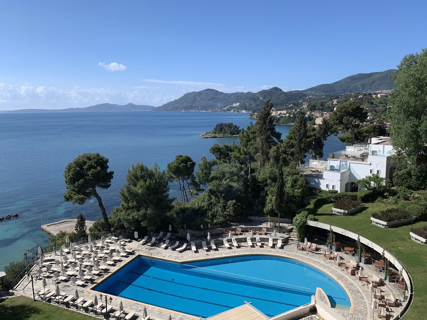Corfu Holiday Palace viesnīcas galvenais baseins Kanoni piekrastē, Korfu salā — skats no augšas uz Jonijas jūru un zaļajiem kalniem tālumā
