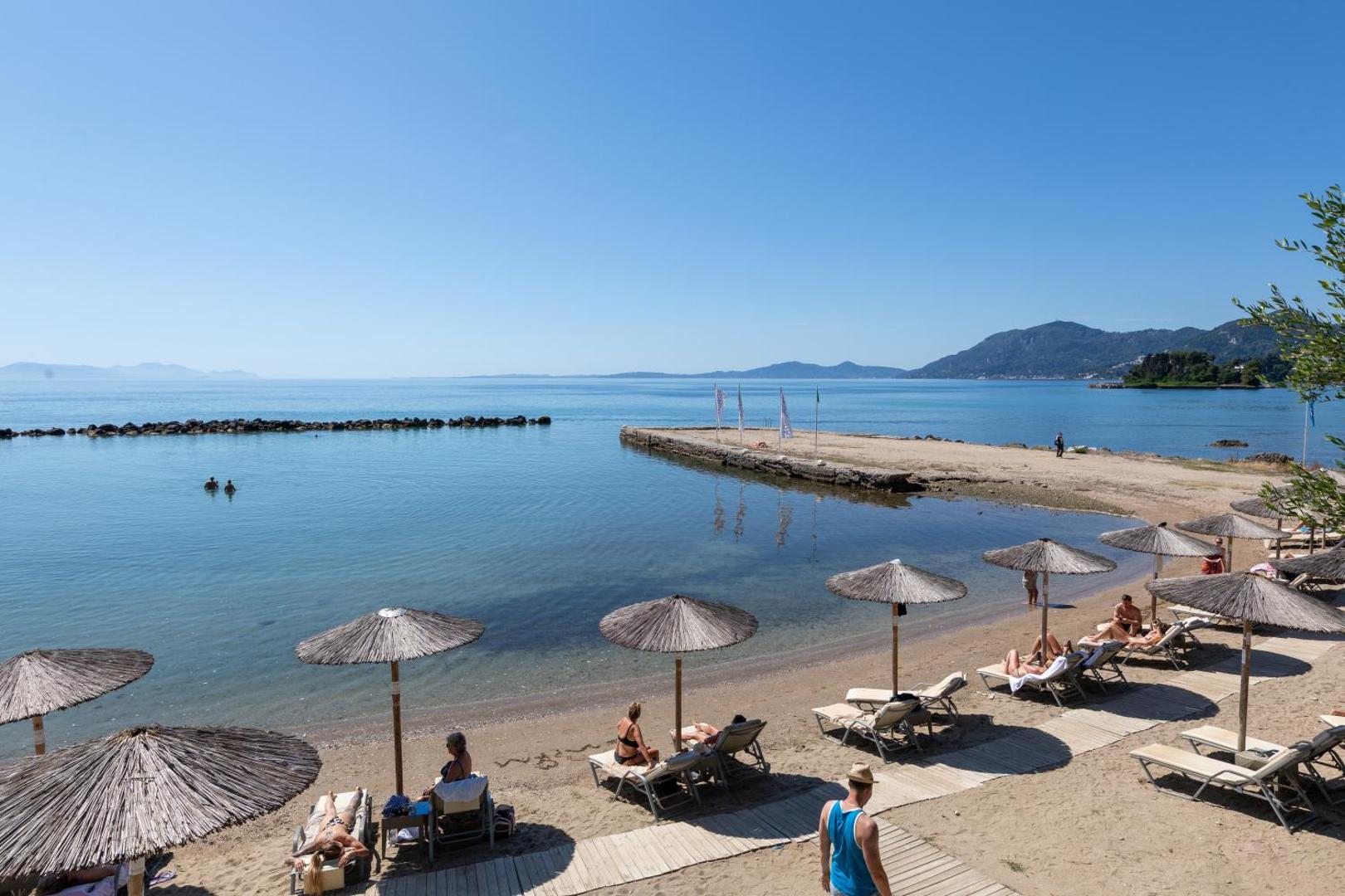Corfu Holiday Palace privātā smilšu pludmale ar salmu saulessargiem, zviļņiem un lēzenu ieeju dzidrā Jonijas jūrā