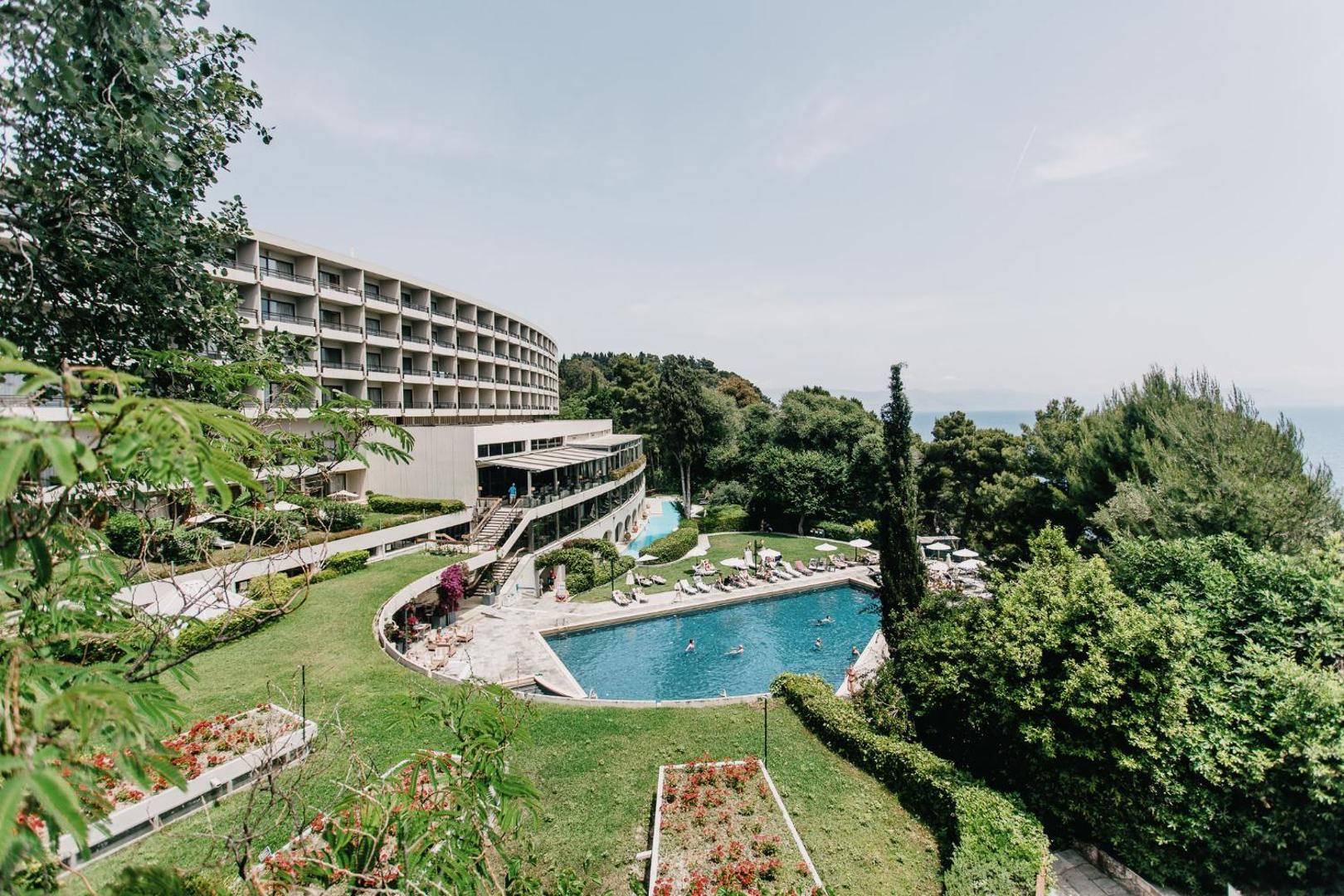 Corfu Holiday Palace galvenā ēka ar balkoniem, āra baseinu un koptu dārzu piekrastē Korfu salā, Grieķijā.