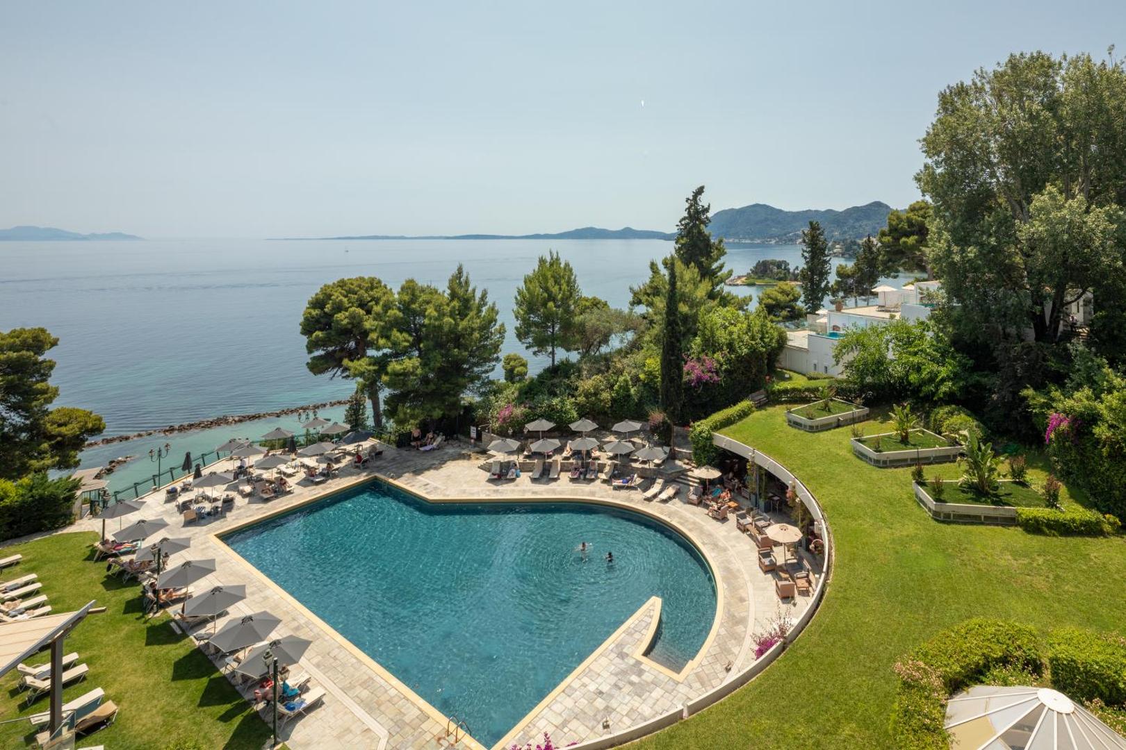 Corfu Holiday Palace āra baseins saulainā dienā ar atpūtas krēsliem, saulessargiem un koptu zālienu Korfu piekrastē, Grieķijā