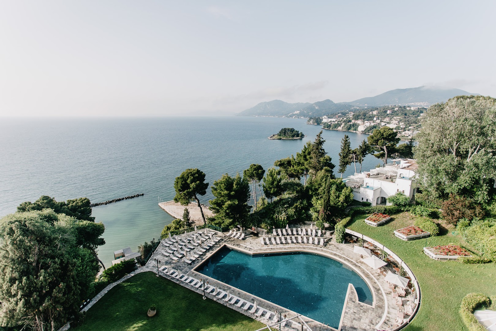 Corfu Holiday Palace skats no augšas uz āra baseinu, piekrasti un Jonijas jūru Korfu salā, Grieķijā — apkārtnē zaļojoši koki.