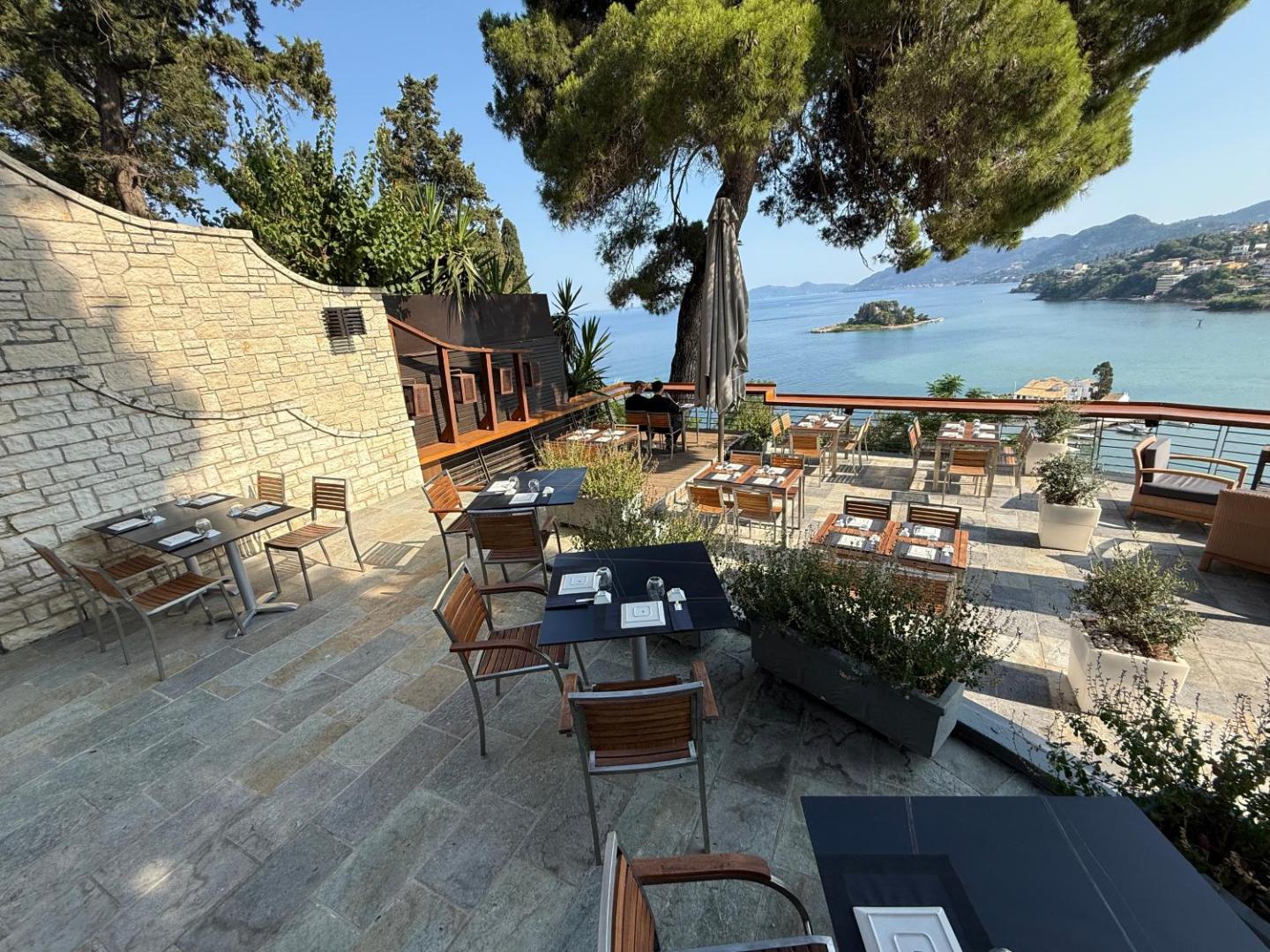 Corfu Holiday Palace āra terases restorāns ar skatu uz Pontikonisi salu Korfu — koka mēbeles un akmens sienas piekrastē