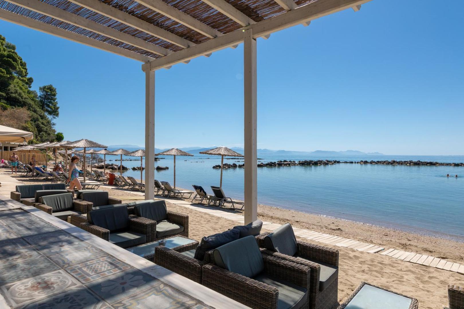 Pludmales bāra terase viesnīcā Corfu Holiday Palace ar ērtiem atpūtas krēsliem un skatu uz jūru zem nojumes
