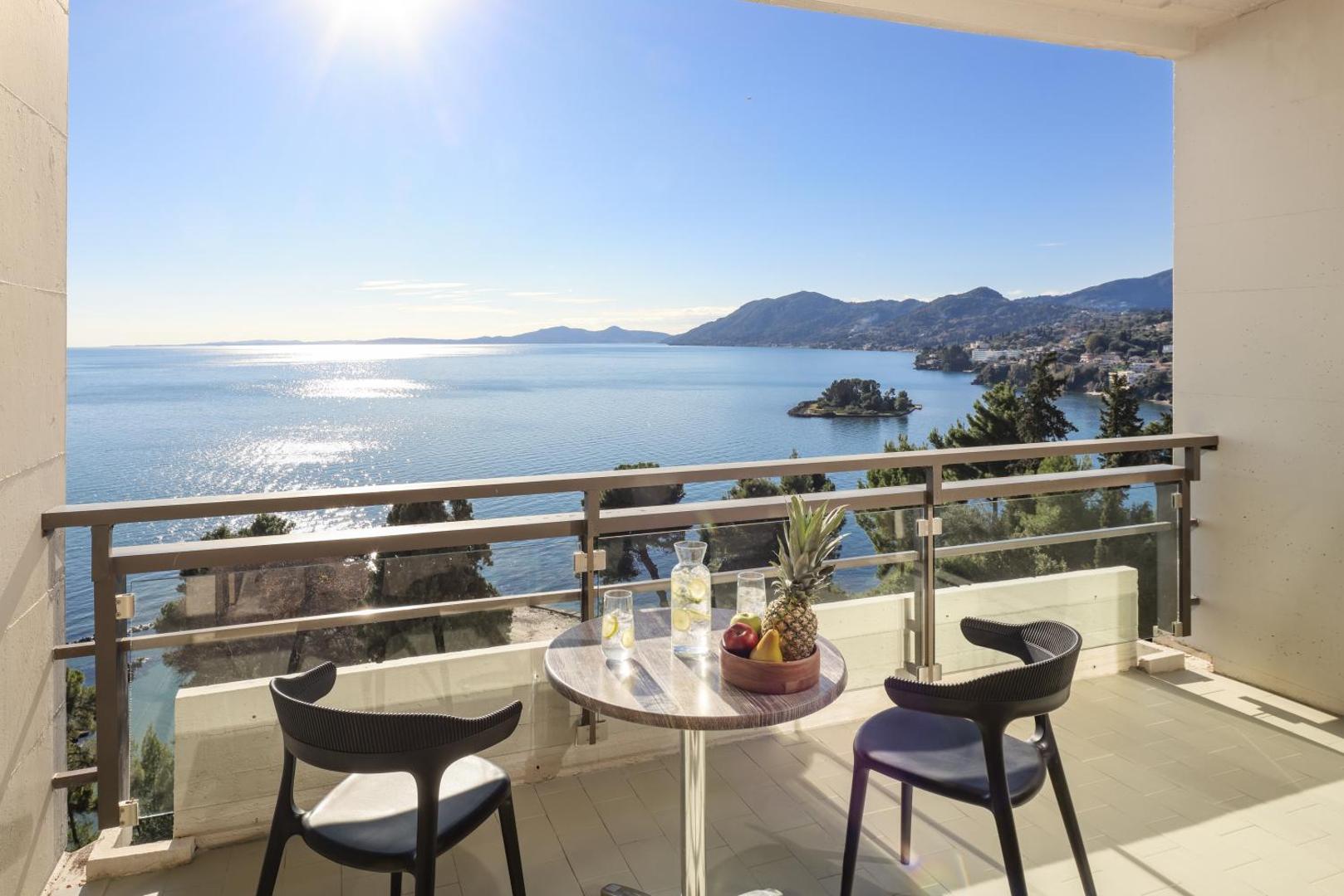 Skats no Corfu Holiday Palace numura balkona uz jūru un kalniem saulainā dienā Korfu salā ar galdiņu un krēsliem