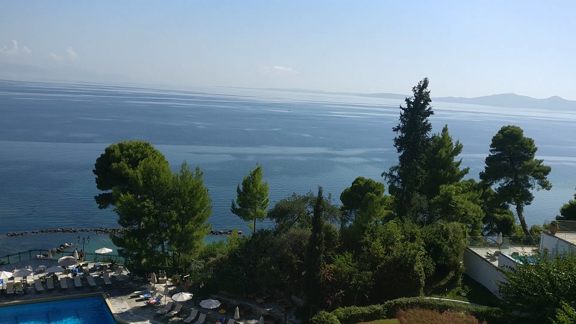 Corfu Holiday Palace āra baseins ar sauļošanās krēsliem un skatu uz Jonijas jūru caur priežu galotnēm
