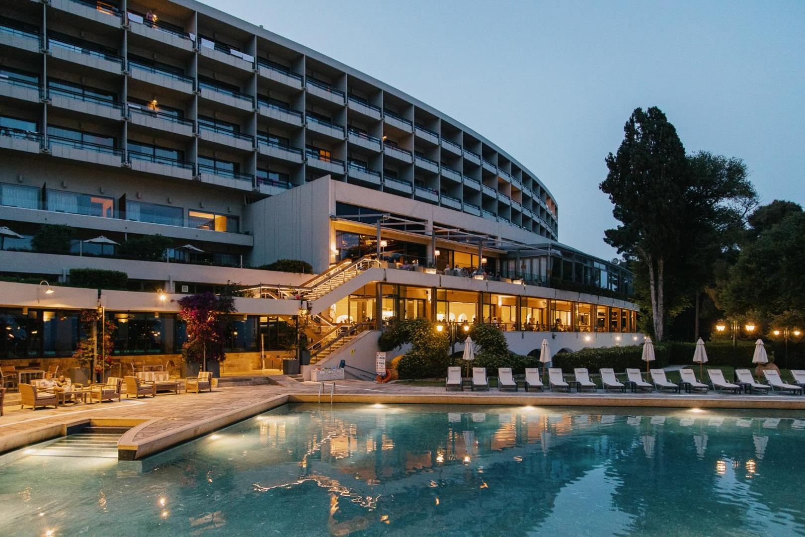 Corfu Holiday Palace viesnīcas fasāde un izgaismots āra baseins vakarā, terase ar atpūtas krēsliem Korfu salā.