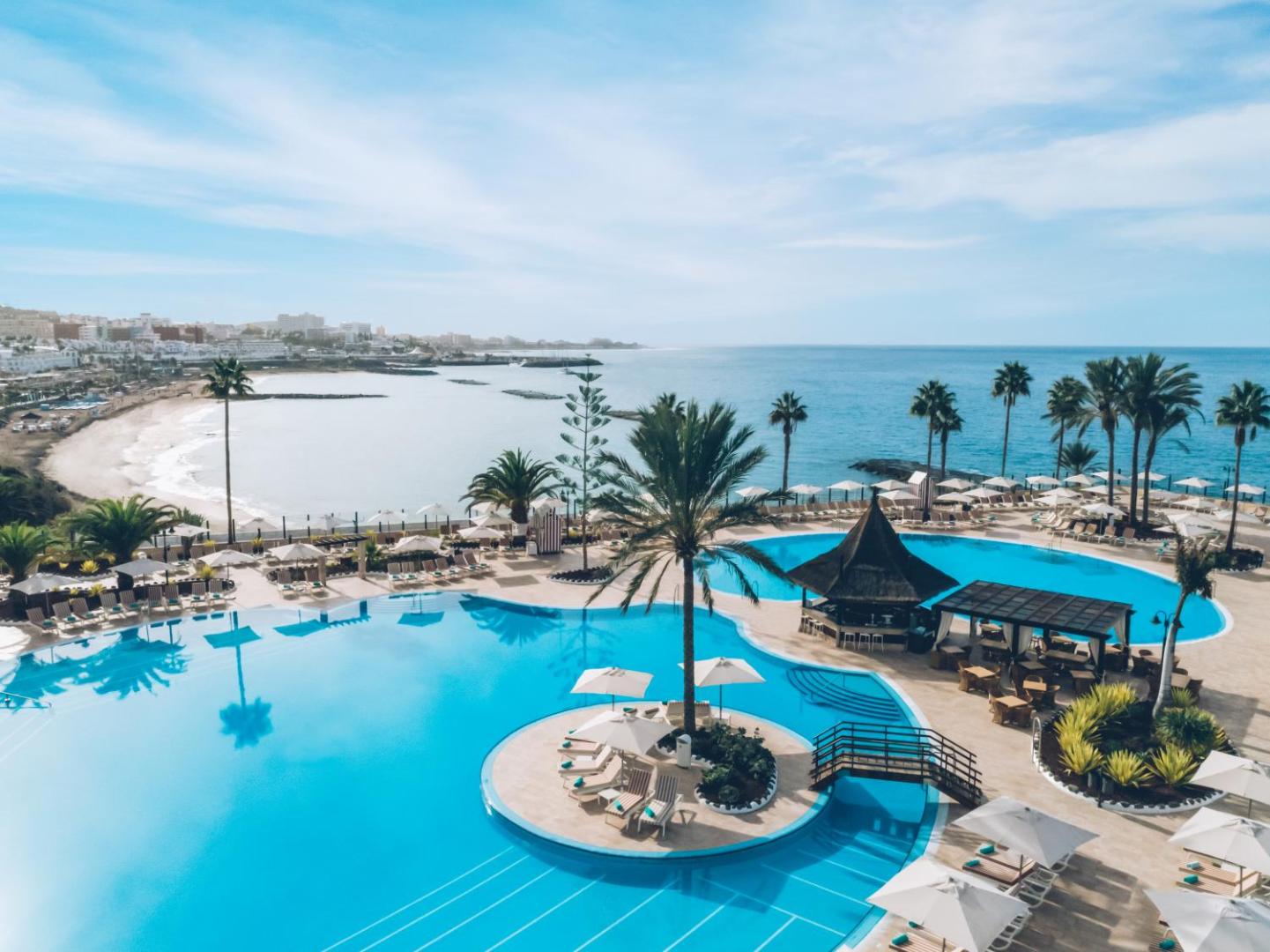 Iberostar Selection Anthelia plašais āra baseinu komplekss pie okeāna Tenerifē ar salu, tiltiņu, bāru un atpūtas krēsliem.
