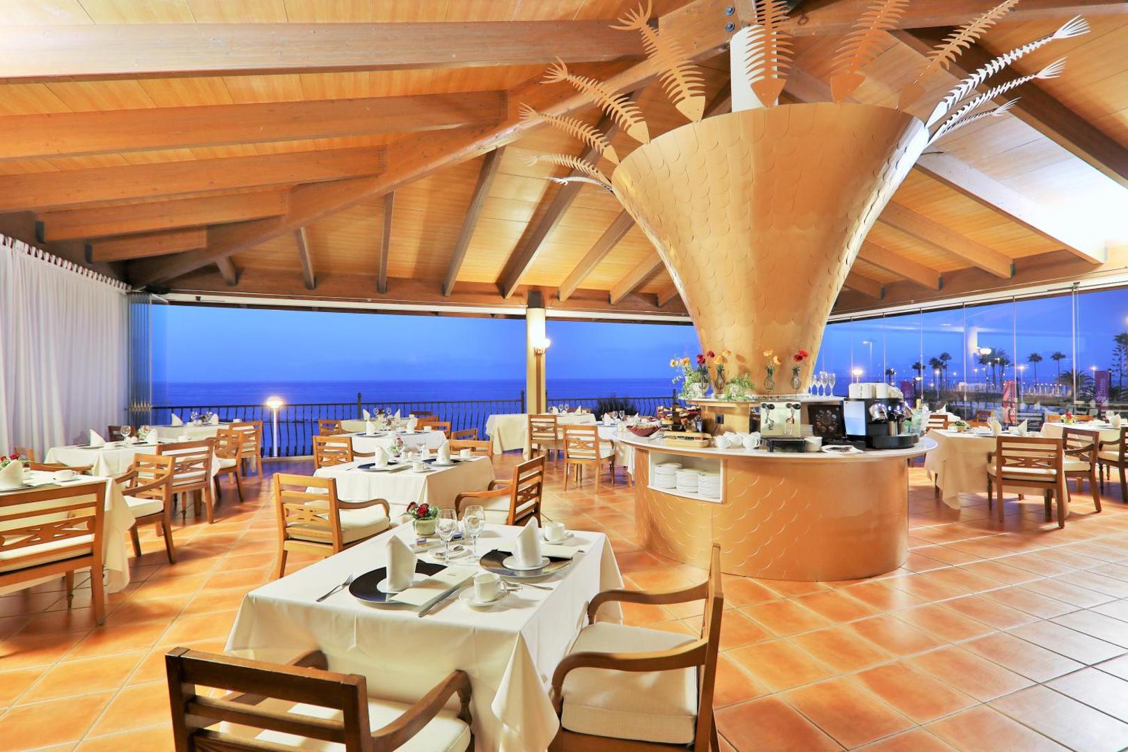 Iberostar Selection Anthelia restorāns ar skatu uz okeānu Tenerifē — eleganti klāti galdi, koka griesti un dekoratīva bufetes zona vakarā.