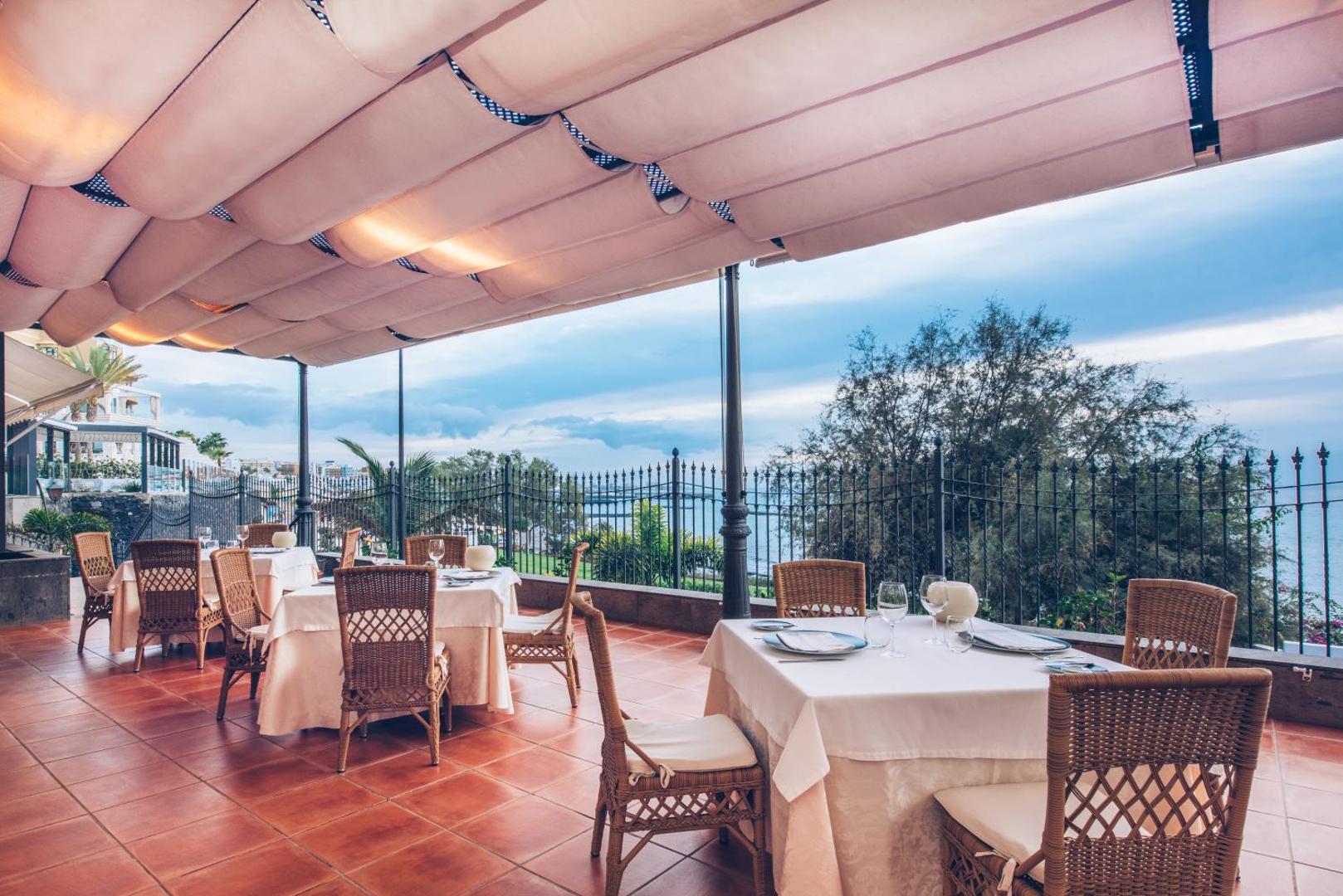 Viesnīcas Iberostar Selection Anthelia restorāna terase ar pītiem krēsliem zem nojumes, skats uz dārzu un okeānu krēslas stundā.