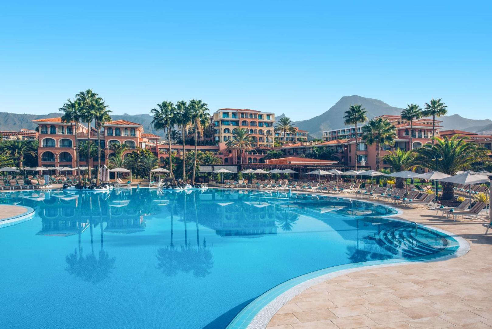 Iberostar Selection Anthelia viesnīcas plašais āra baseins Tenerifē, Spānijā — fonā redzama viesnīcas arhitektūra, palmas un kalnu ainava saulainā dienā.