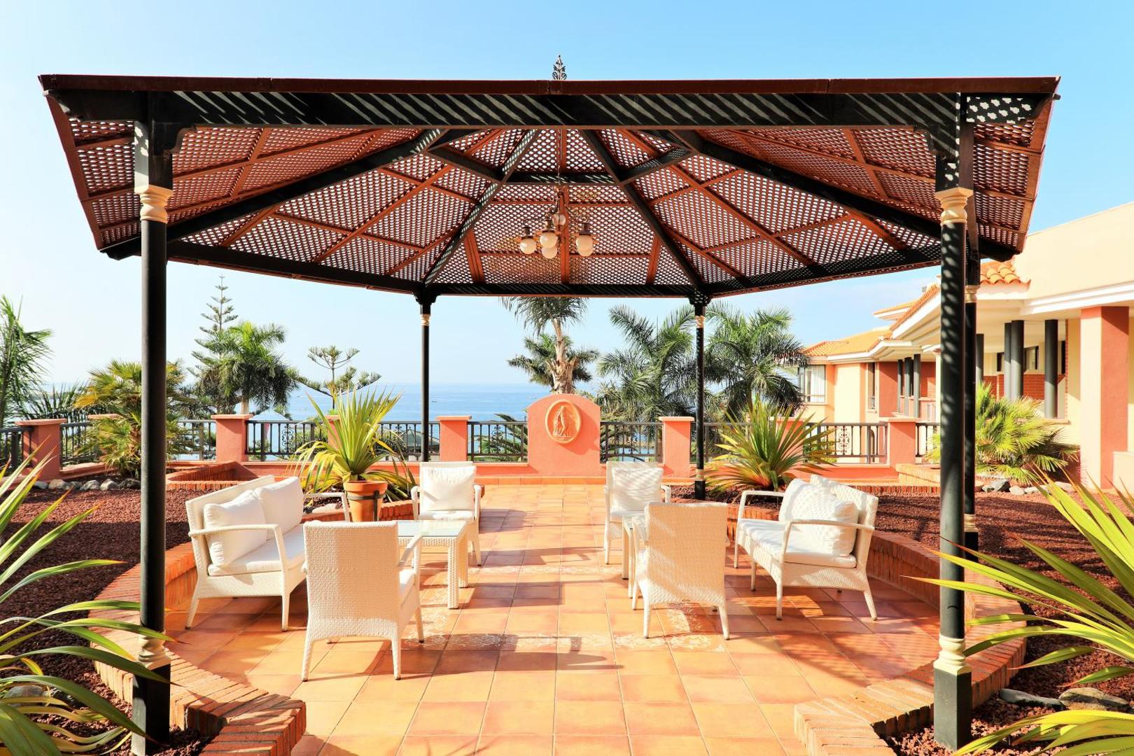 Iberostar Selection Anthelia atpūtas terase ar koka lapeni un baltiem krēsliem Tenerifē, no kuras paveras skats uz okeānu un palmām