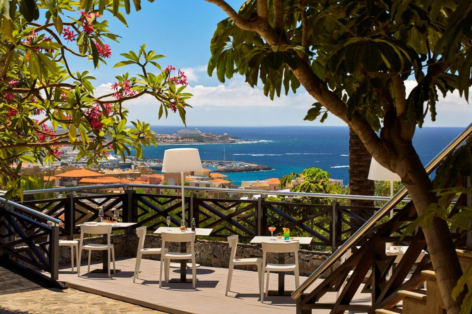 Meliā Jardines del Teide mājīga terase ar ziedošiem kokiem un skatu uz piekrastes pilsētu un okeānu