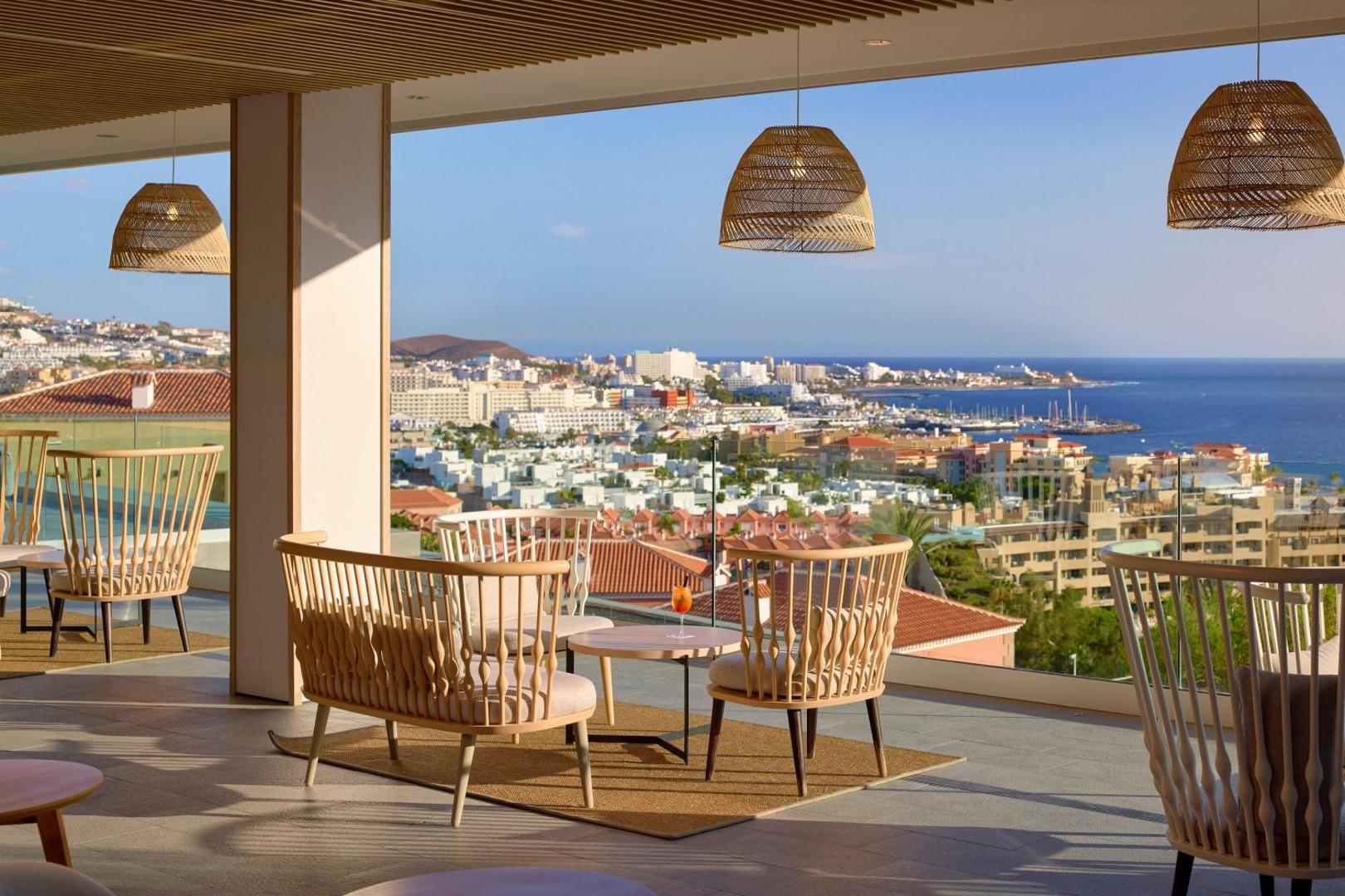 Skats no Melia Jardines del Teide terases uz kūrortpilsētu un okeānu saulrietā, mēbelēta atpūtas zona ar pītiem krēsliem