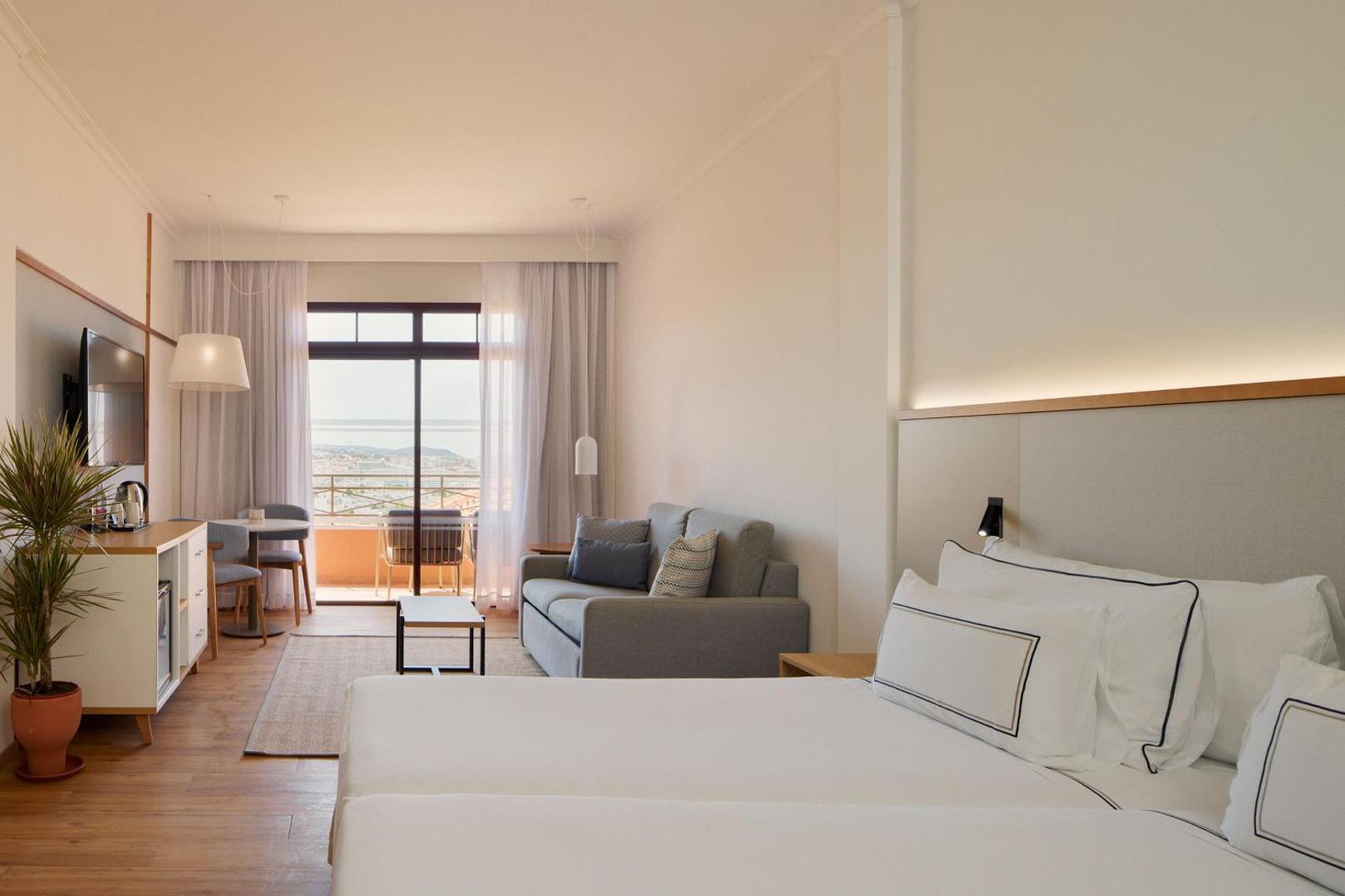 Meliá Jardines del Teide numura interjers ar skatu uz balkonu un okeānu — gaiša guļamistaba ar dīvānu un darba zonu
