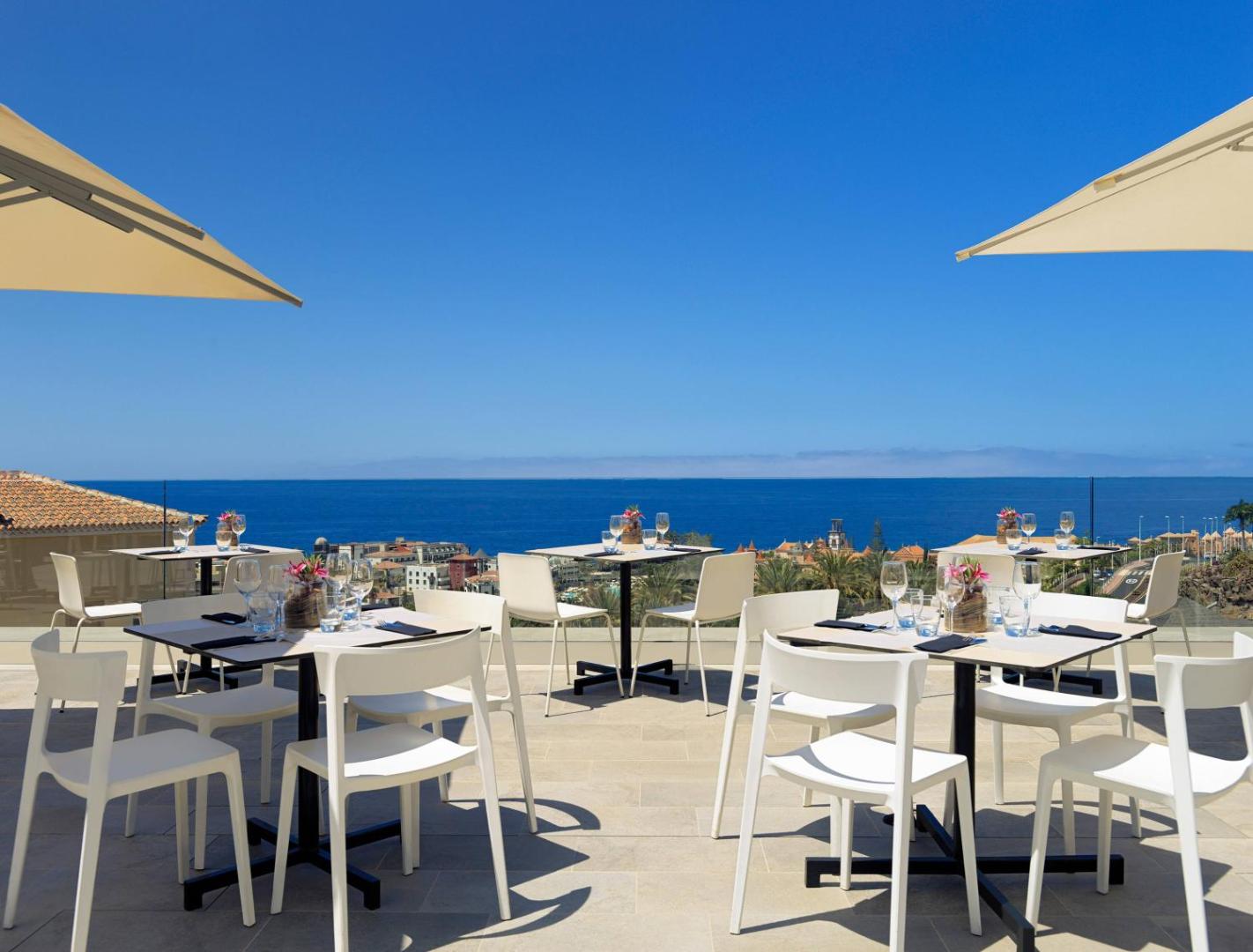 Viesnīcas Meliá Jardines del Teide restorāna āra terase ar baltiem galdiem un skatu uz zilo okeānu