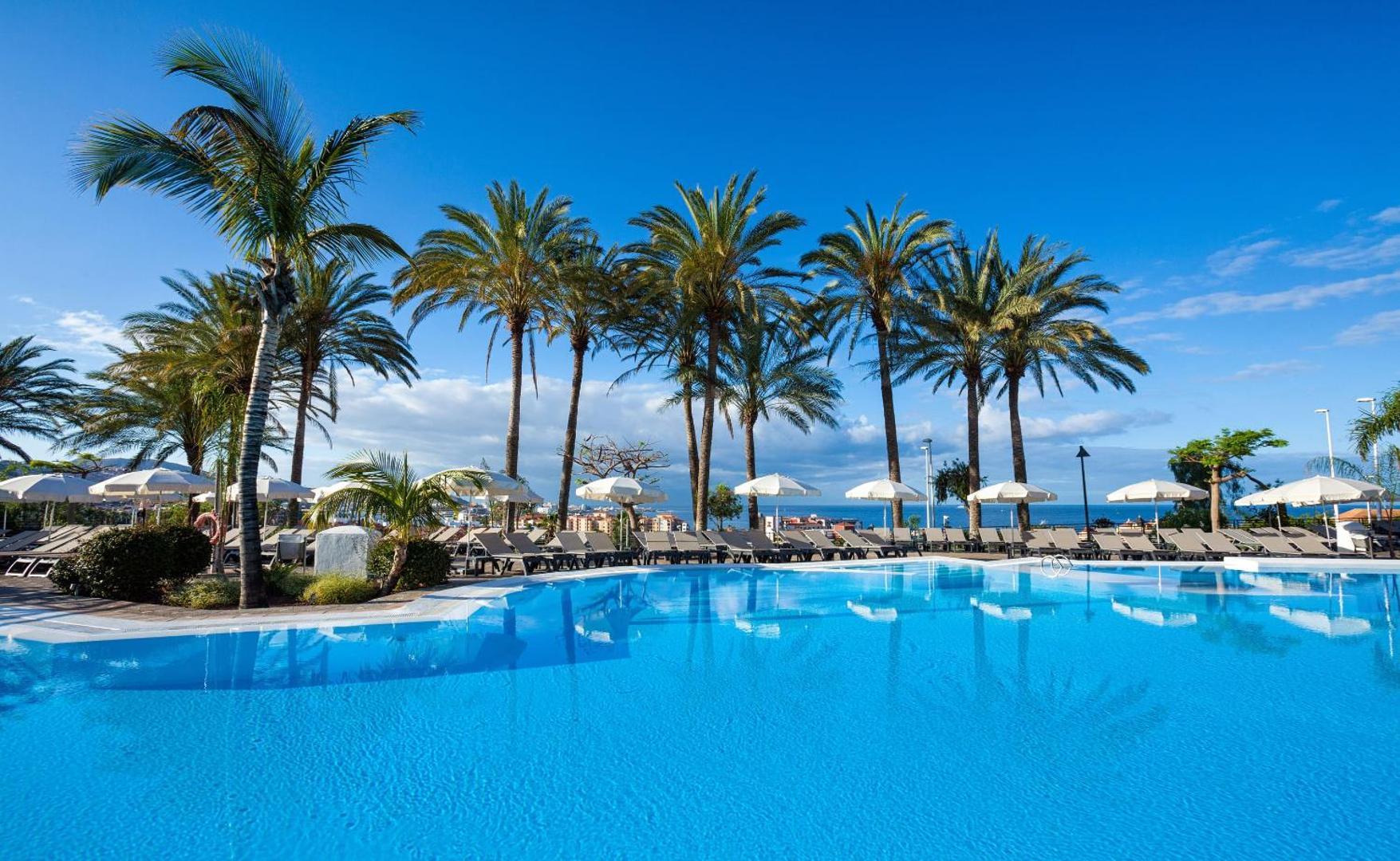 Melia Jardines del Teide āra baseins ar dzidru ūdeni, sauļošanās krēsliem un augstām palmām pret zilu debesu fonu Tenerifē