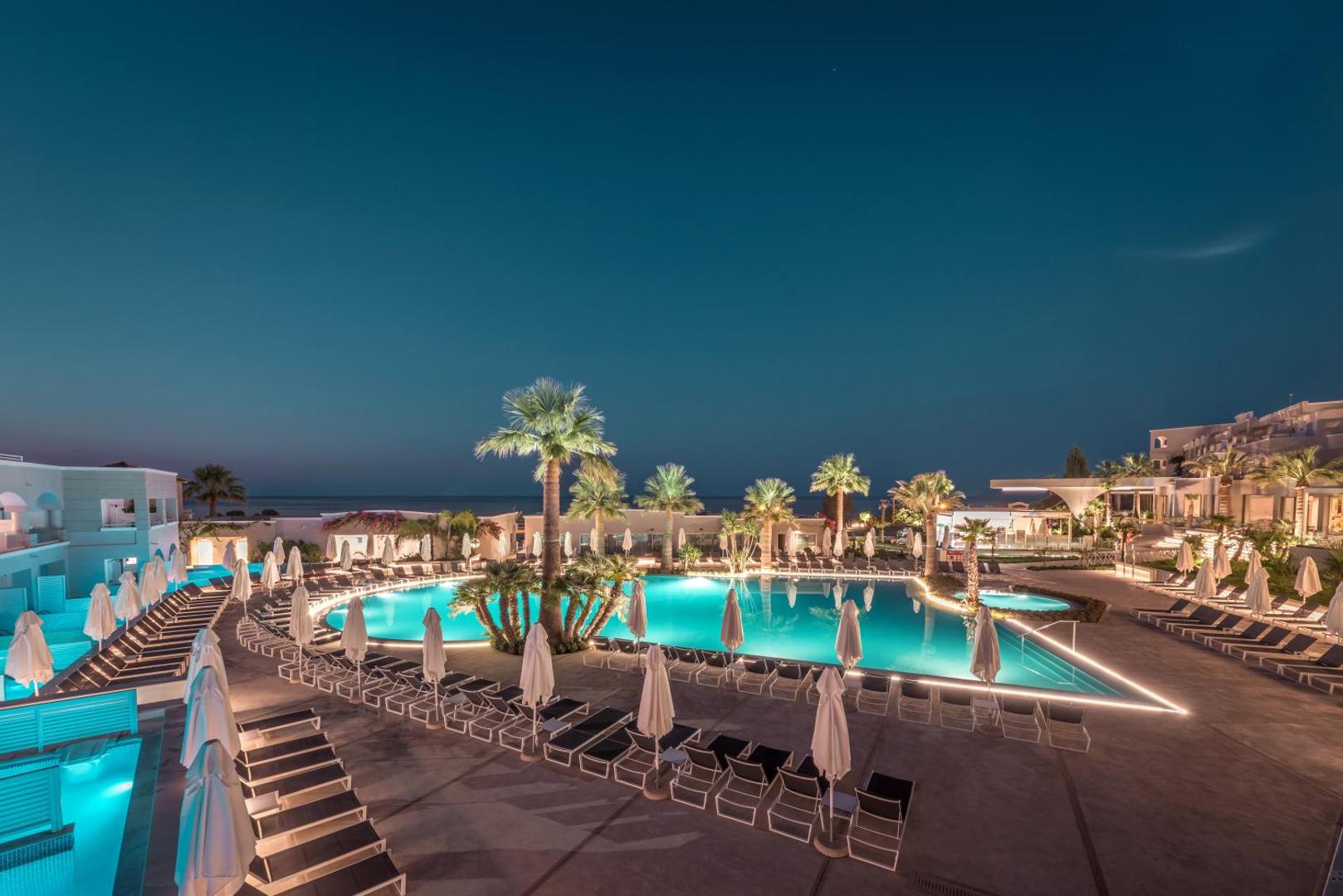 Mitsis Rodos Village viesnīcas galvenais baseins vakarā ar izgaismotu ūdeni, palmām un sauļošanās krēsliem Rodas piekrastē