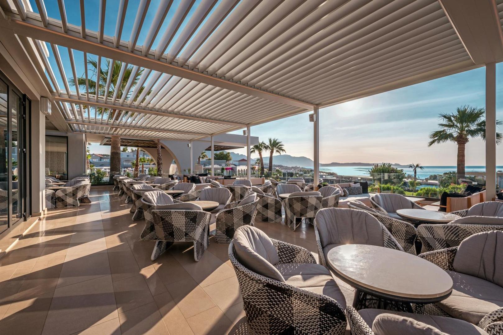 Plaša Mitsis Rodos Village atpūtas terase ar moderniem pītiem krēsliem un panorāmas skatu uz piekrasti saulrietā