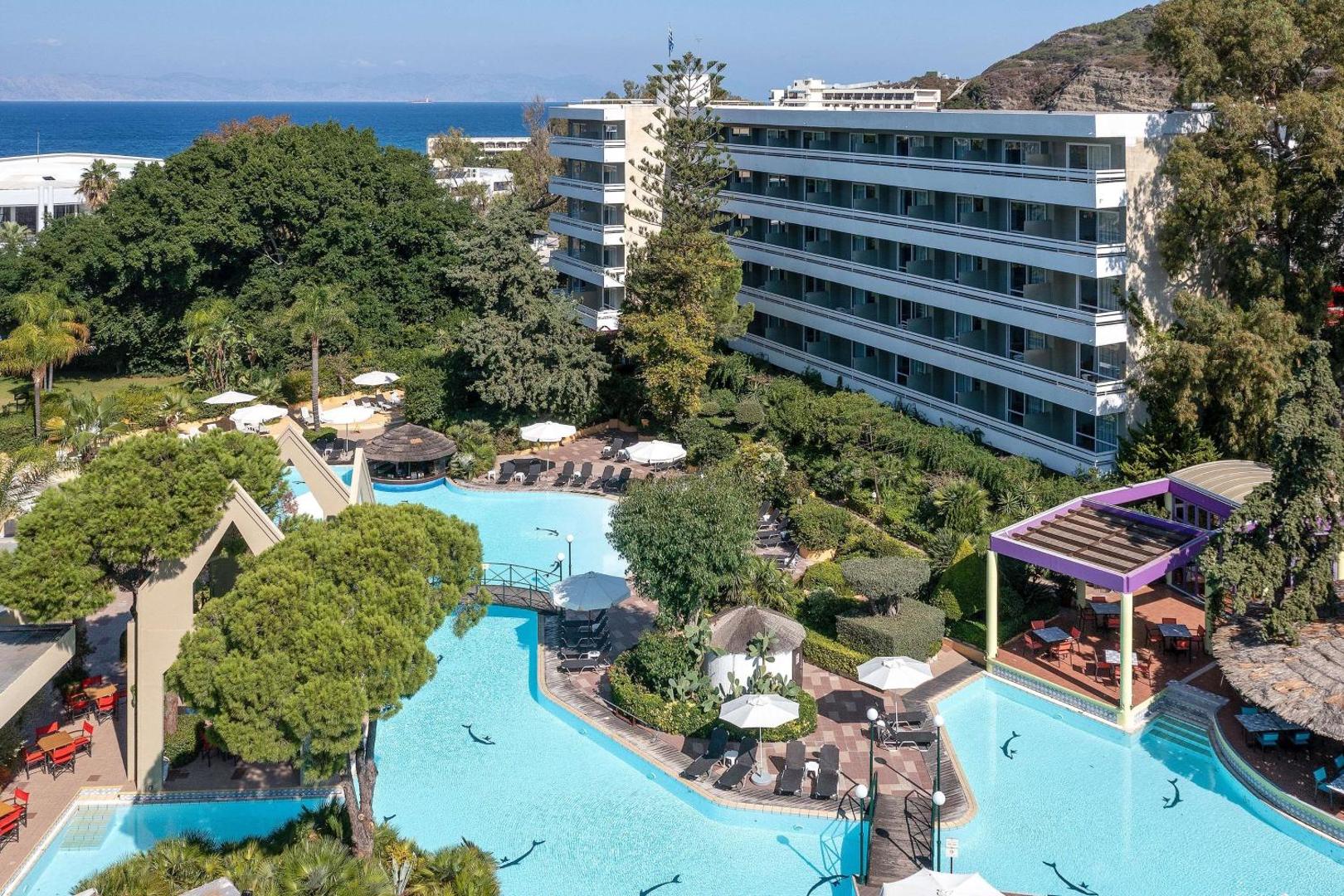 Dionysos Hotel Rodas salā — skats no augšas uz plašo baseinu kompleksu, ko ieskauj zaļojošs dārzs un palmas netālu no jūras krasta