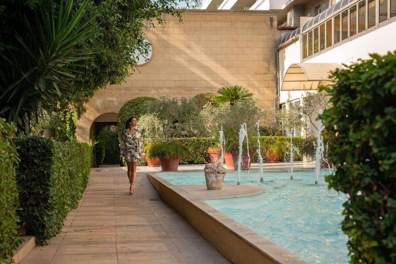 Dionysos Hotel iekšpagalms ar dekoratīvām strūklakām un koptu dārzu, kurā pastaigājas viesis pie akmens sienas ēkas