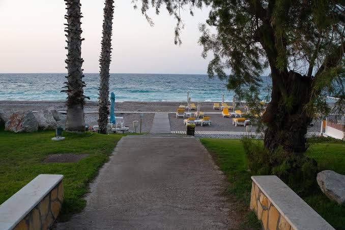 Viesnīcas Dionysos pieeja oļu pludmalei Rodas salā — skats no dārza uz jūru ar sauļošanās krēsliem un palmām