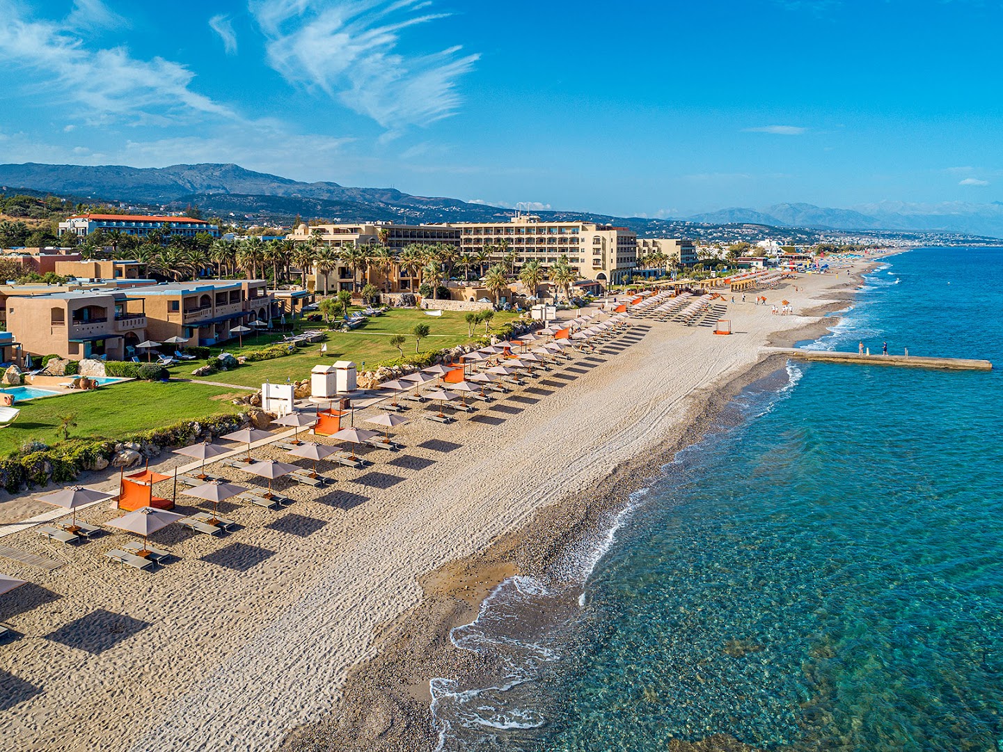 Aquila Rithymna Beach viesnīcas plašā smilšu pludmale Krētā ar saulessargiem, zvilnēm un skatu uz kūrorta ēkām un kalniem tālumā