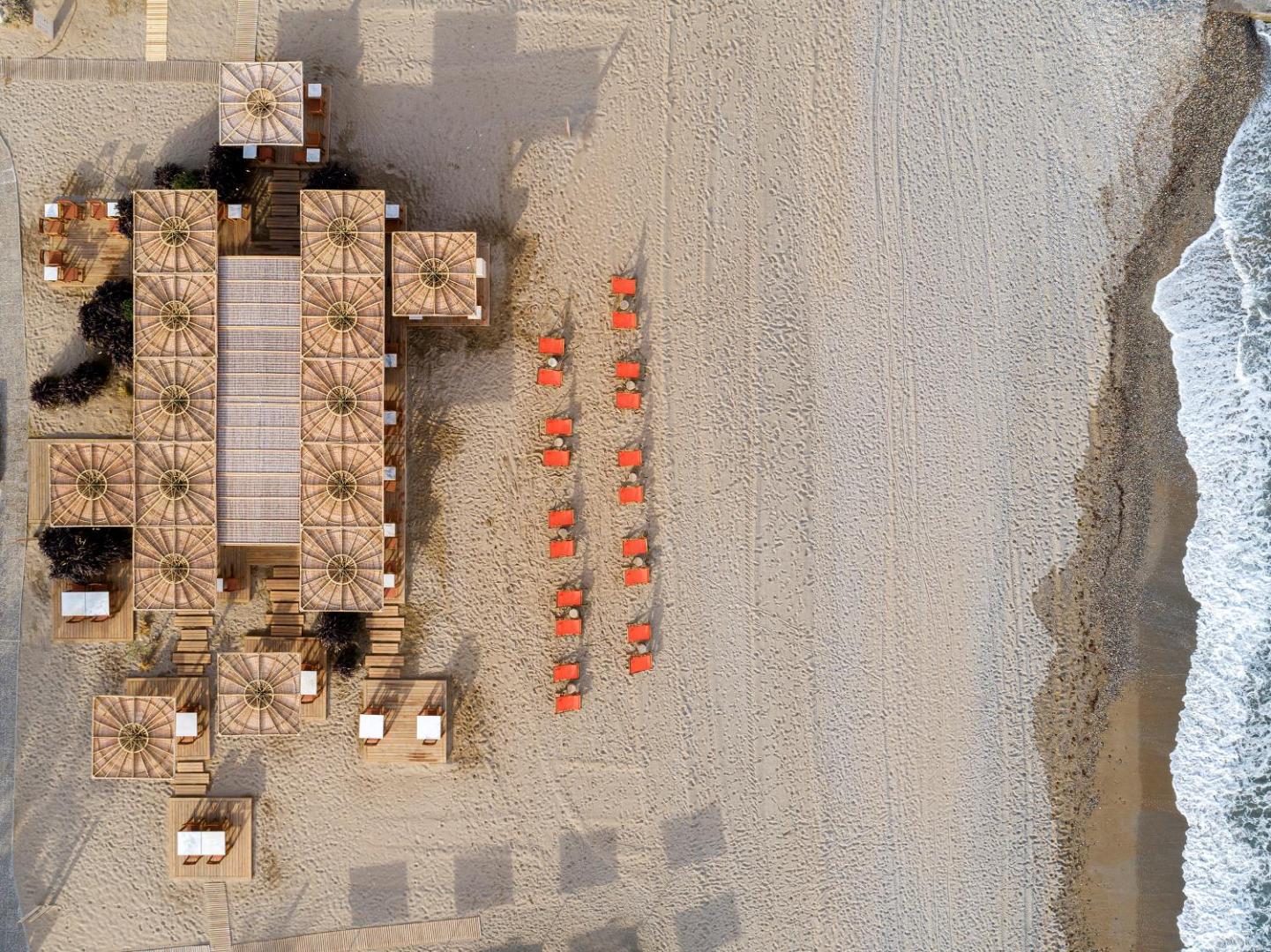 Aquila Rithymna Beach viesnīcas pludmales bāra un atpūtas zonas skats no putna lidojuma Krētā ar stilīgām koka konstrukcijām un oranžiem sauļošanās krēsliem.