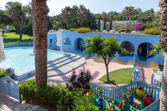 Aquila Rithymna Beach bērnu baseins un rotaļu laukums — zila ēka ar arkām, sekls baseins un zaļa teritorija