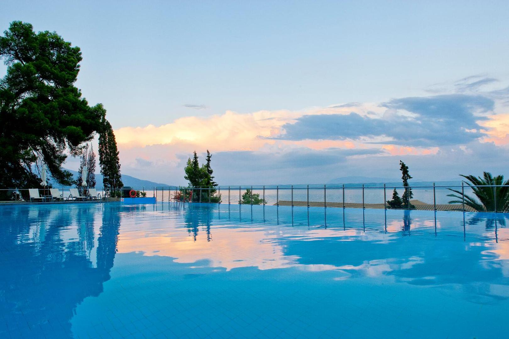 Kontokali Bay Resort & Spa baseins saulrieta laikā ar debesu atspulgu ūdenī un skatu uz piekrasti Korfu