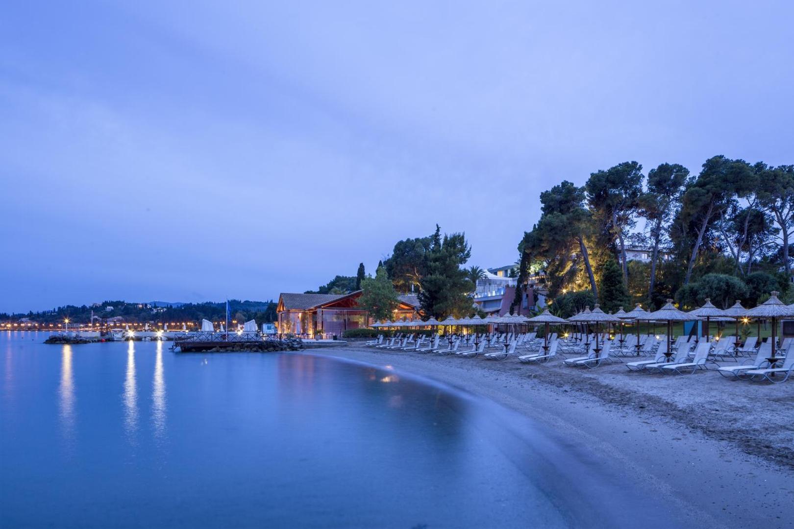 Kontokali Bay Resort & Spa privātā pludmale vakarā ar saulessargiem, zviļņiem un izgaismotu piekrastes restorānu Korfu
