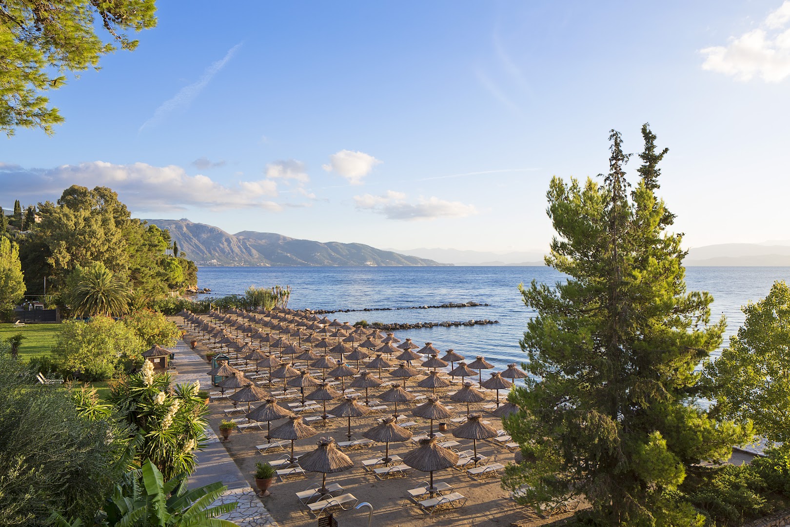 Kontokali Bay Resort & Spa privātā pludmale Korfu salā ar salmu saulessargiem, zviļņiem un skatu uz Jonijas jūru un kalniem