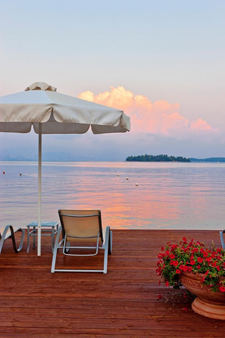 Saulrieta skats no Kontokali Bay Resort & Spa koka terases ar sauļošanās krēslu un saulessargu virs Jonijas jūras