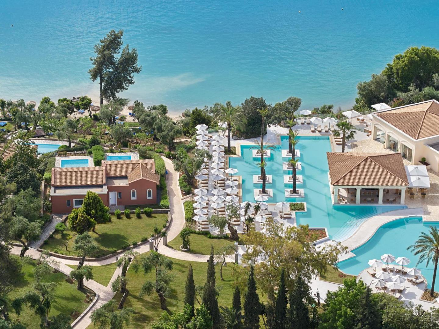 Grecotel Eva Palace teritorijas skats no augšas, parādot baseinu kompleksu, bungalo tipa ēkas un tiešu piekļuvi pludmalei.