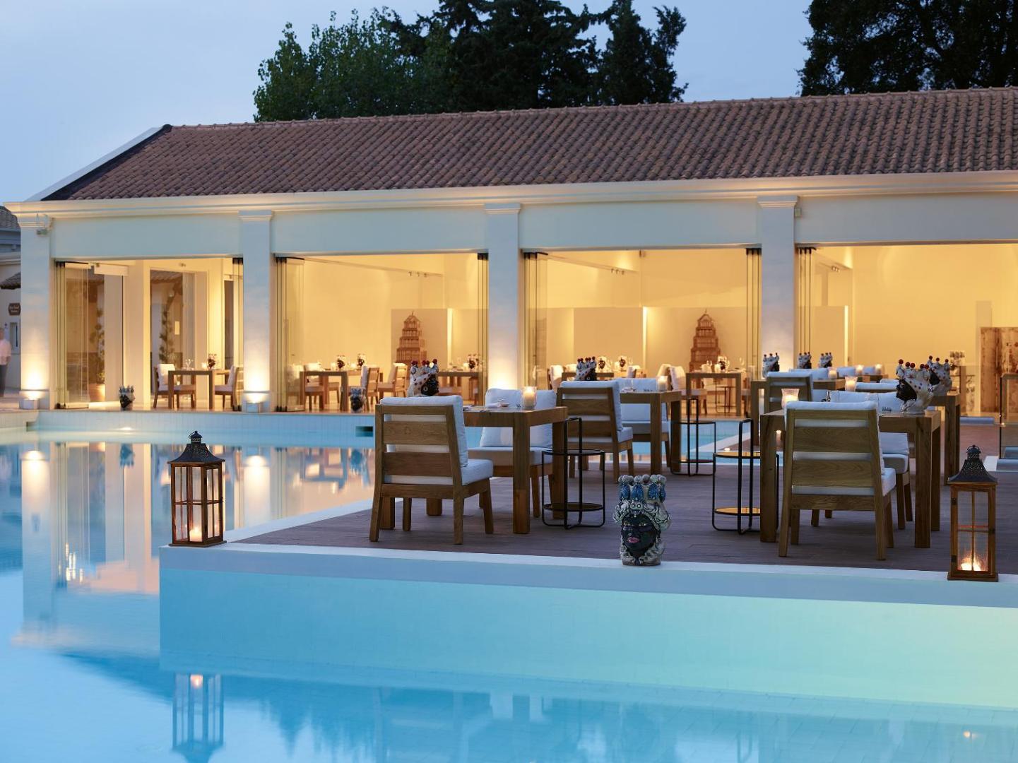 Grecotel Eva Palace vakara apgaismojums pie baseina un restorāna terases ar klātiem galdiem un dekoratīvām laternām