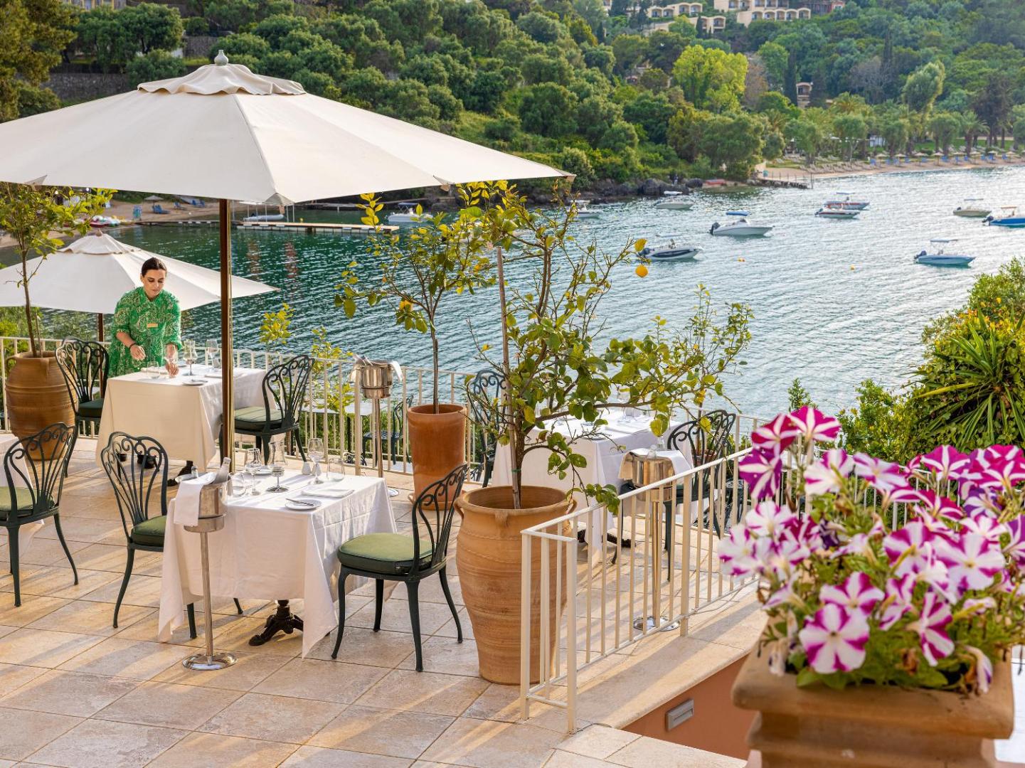 Grecotel Eva Palace restorāna āra terase ar skatu uz Jonijas jūru Korfu salā — balti klāti galdi un citrona koki podos