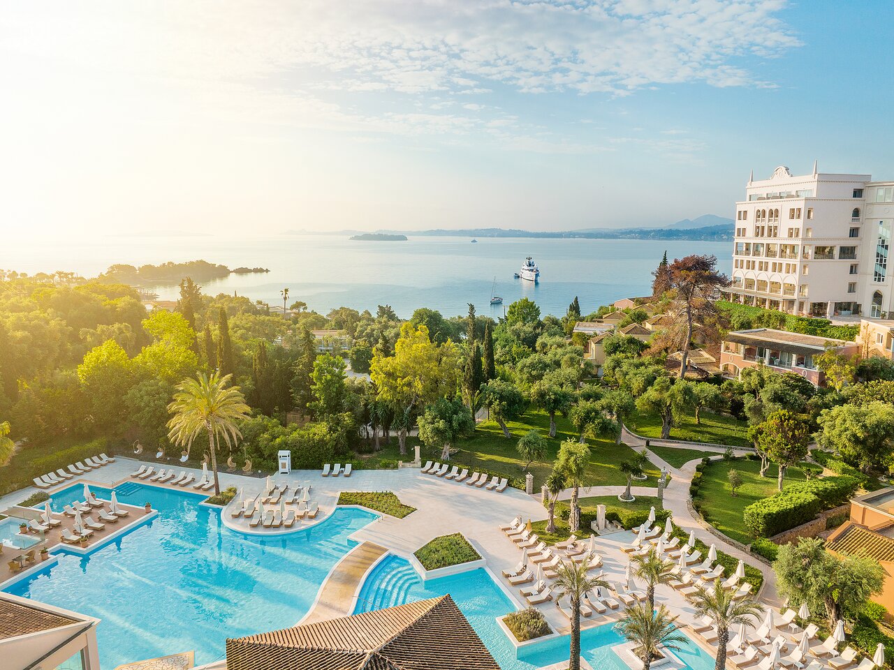 Grecotel Eva Palace panorāmas skats uz galveno baseinu, zaļojošu dārzu un Jonijas jūru Korfu piekrastē saulrieta gaismā.