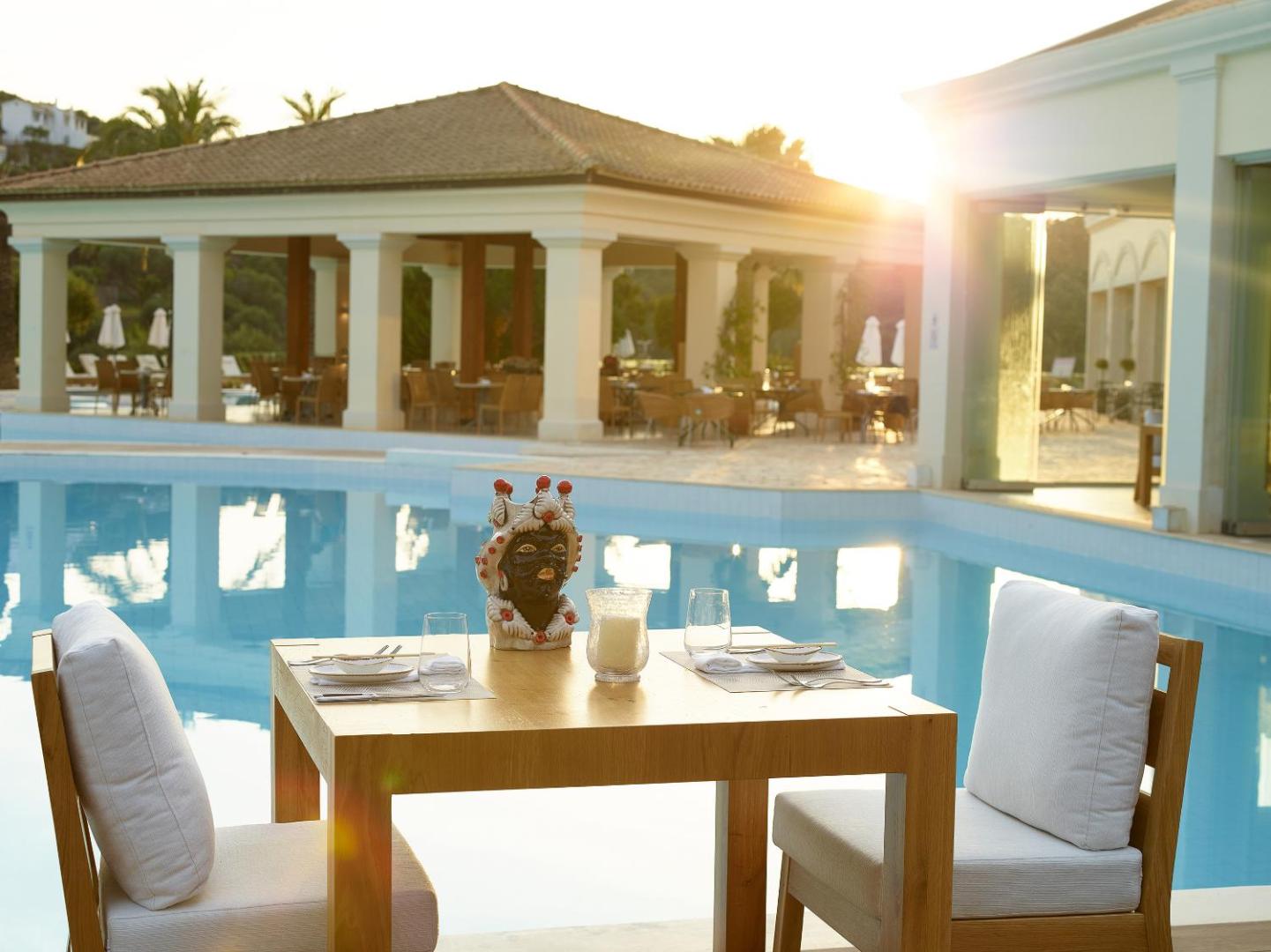 Grecotel Eva Palace eleganti klāts galds divām personām pie āra baseina saulrieta gaismā ar skatu uz restorānu
