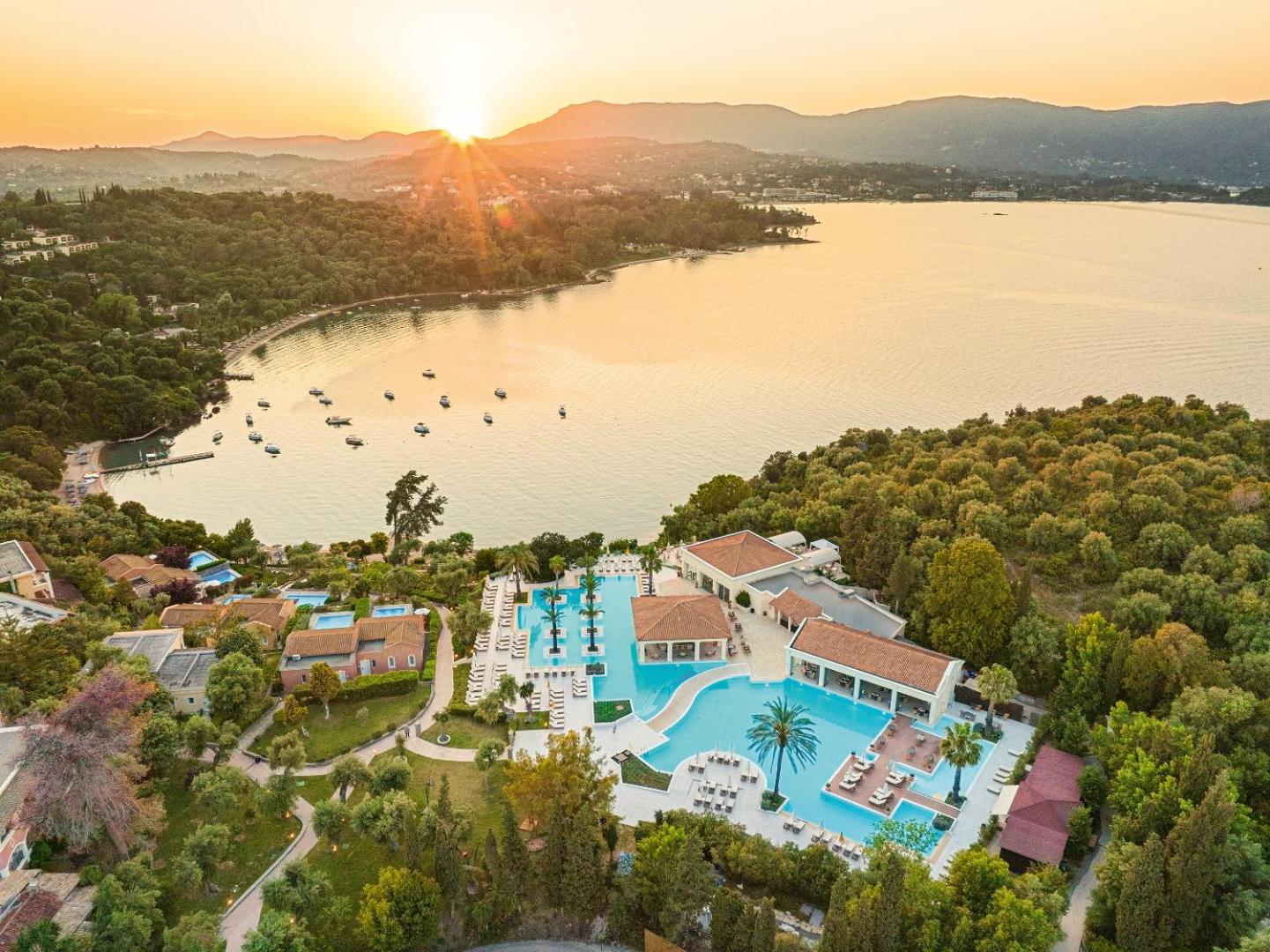 Saulrieta skats pār Grecotel Eva Palace baseiniem un līci Korfu — zeltaina gaisma apspīd kalnus un pietauvotās laivas.