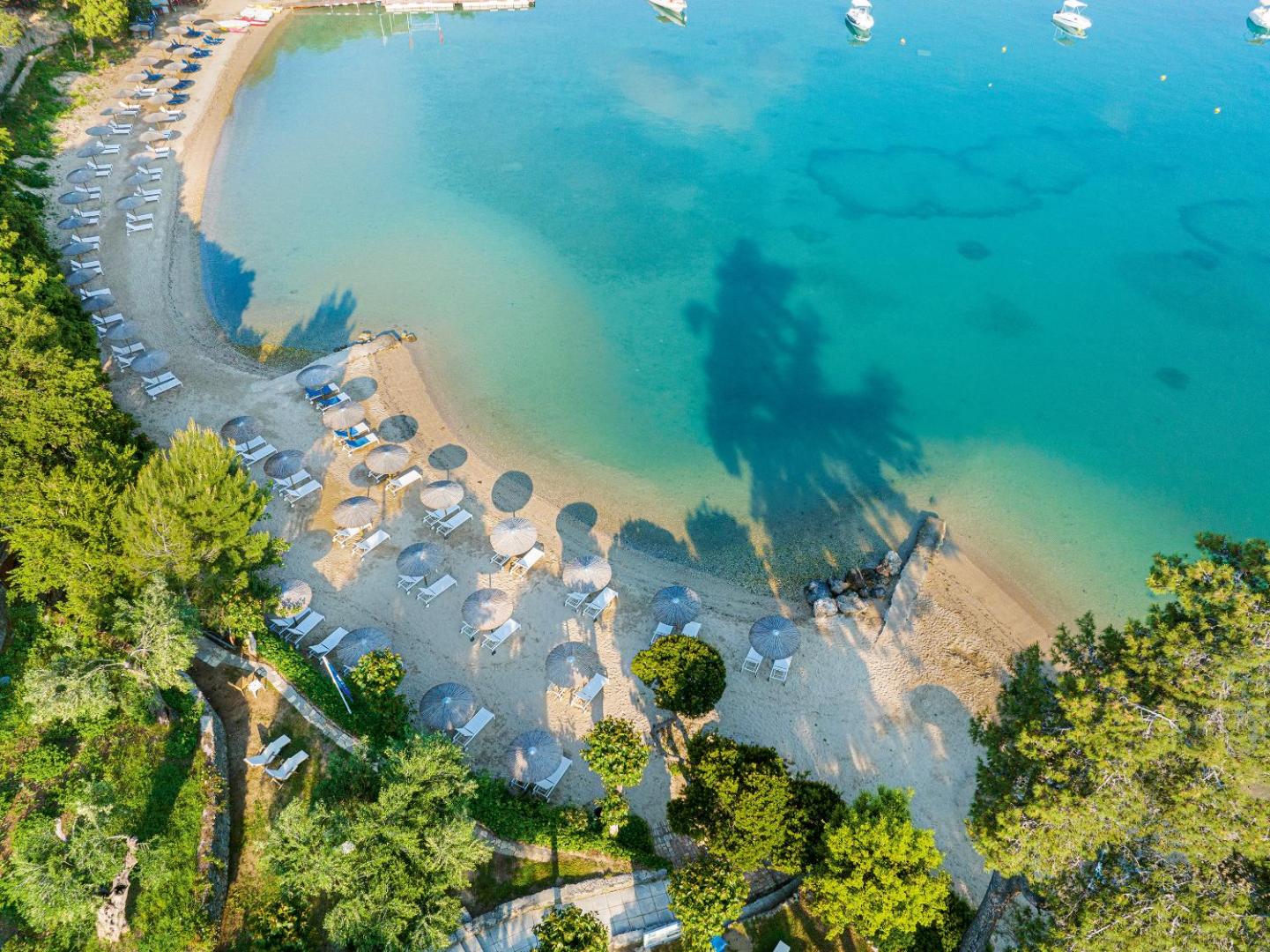 Grecotel Eva Palace privātā pludmale Korfu no putna lidojuma — tirkīzzils ūdens, smilšu krasts un saulessargi koku ieskāvumā