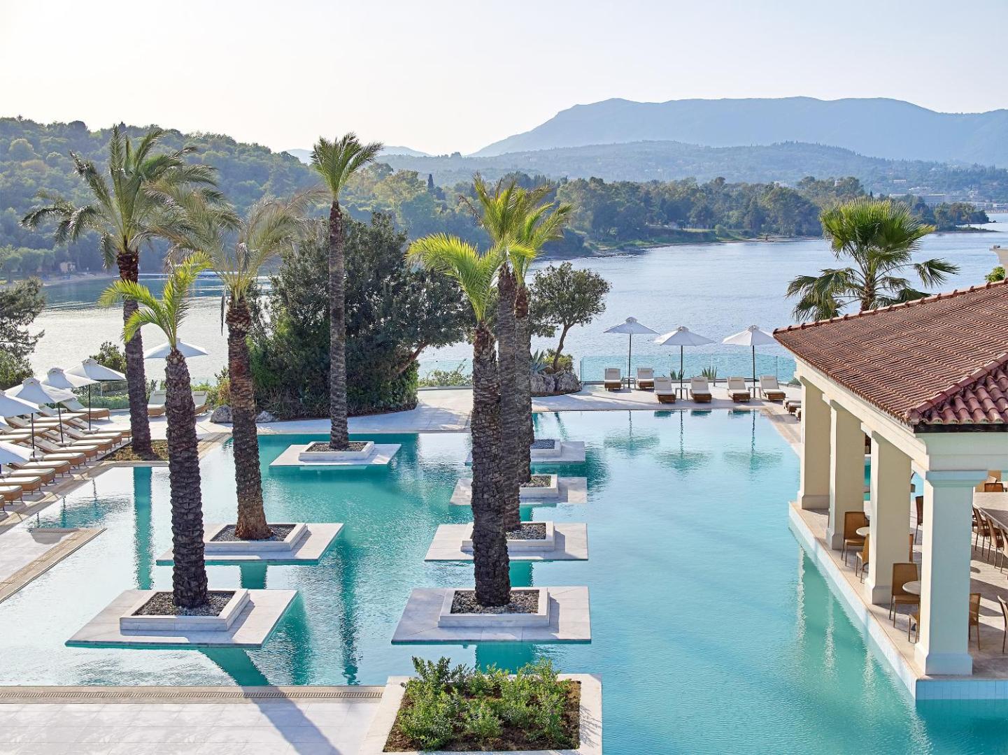 Grecotel Eva Palace baseins ar palmām un skatu uz kalniem un jūru, saulessargi un atpūtas krēsli pie ūdens.