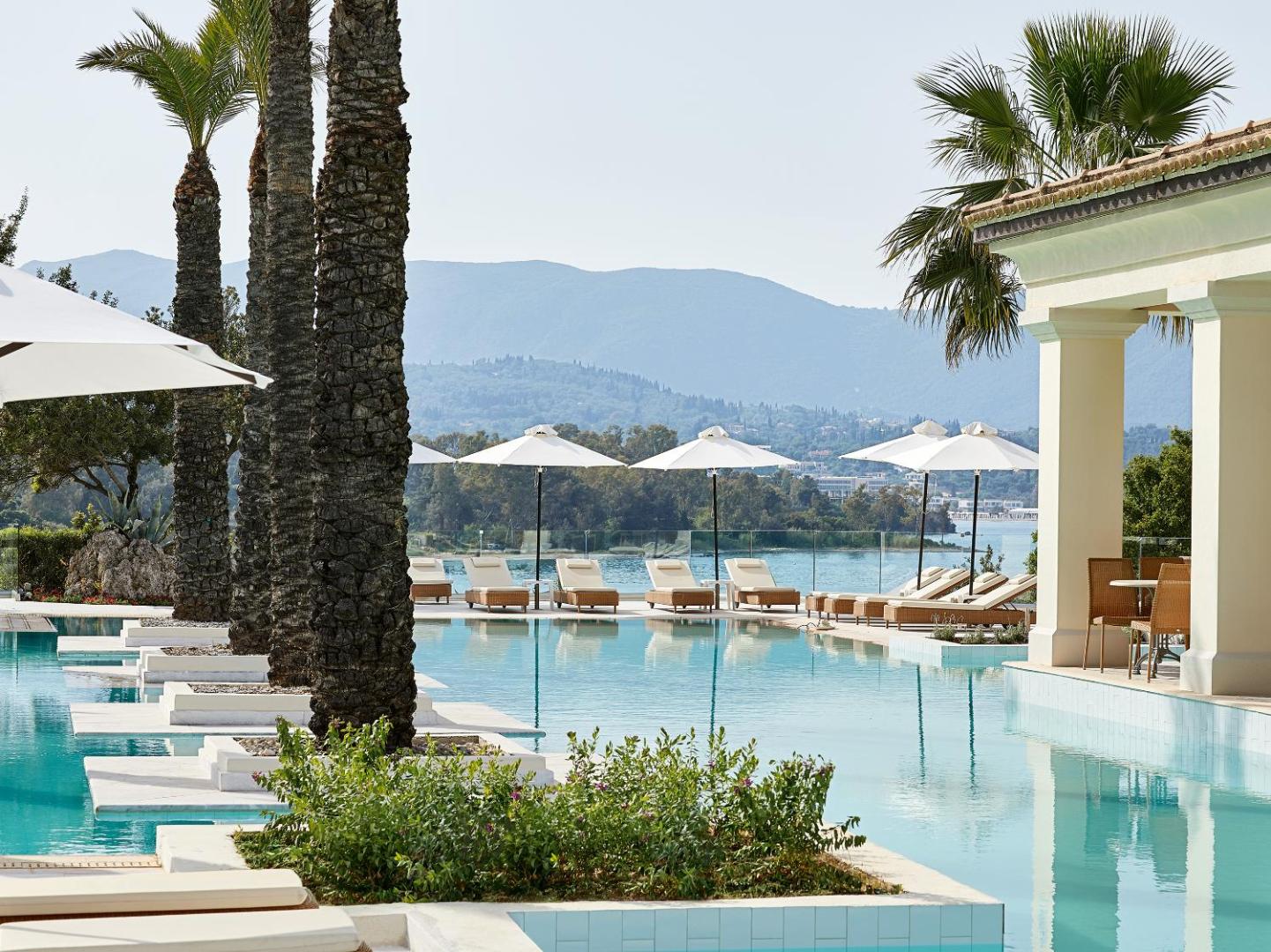 Grecotel Eva Palace baseina mala ar elegantiem atpūtas krēsliem, baltiem saulessargiem un skatu uz piekrastes kalniem.
