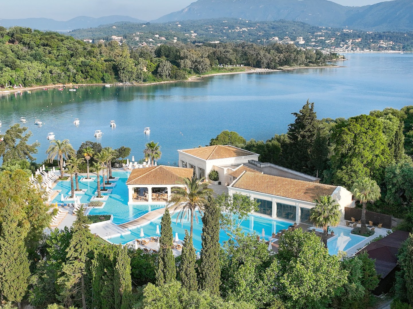 Grecotel Eva Palace baseina zona ar bāru un palmām, fonā kalnainā Korfu ainava un mierīgs jūras līcis.