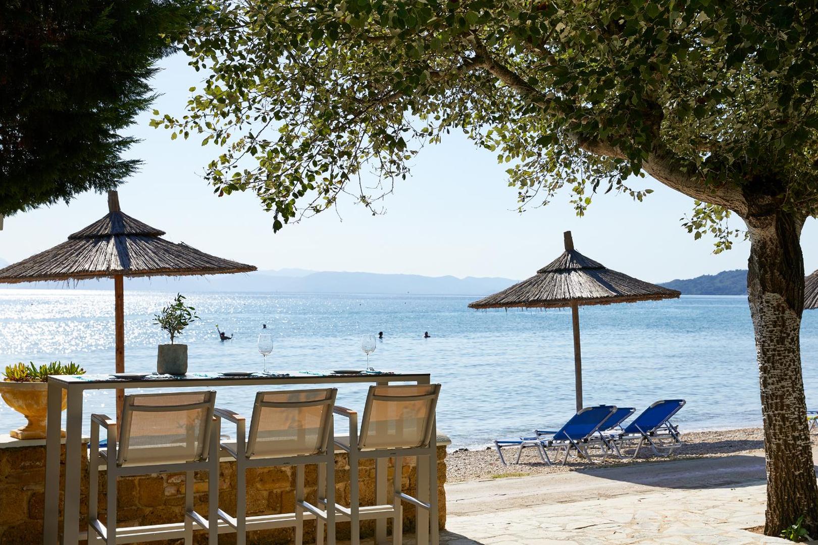 Delfinia Hotel bāra zona pie pludmales Korfu salā ar skatu uz Jonijas jūru, galdiem un saulessargiem koku pavēnī