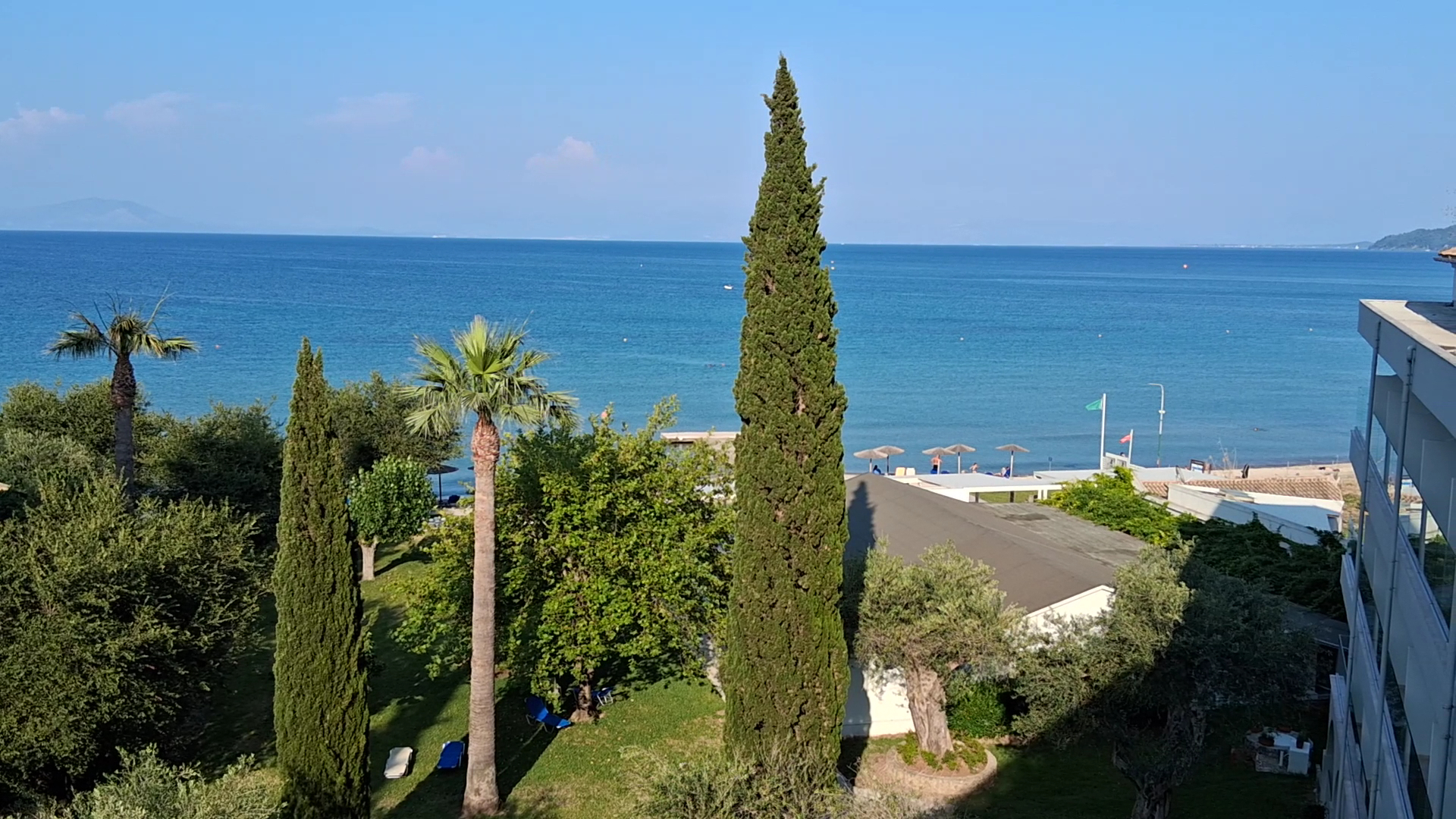 Skats uz Jonijas jūru no Delfinia Hotel teritorijas Korfu — augstas cipreses un palmas dārzā pie pašas pludmales