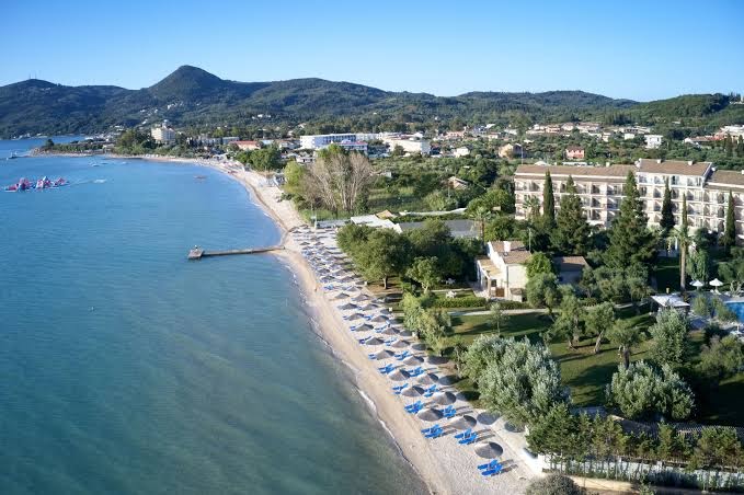 Delfinia Hotel pludmales līnija no augšas — smilšu un oļu krasts ar saulessargiem, guļamkrēsliem un skatu uz Jonijas jūras līci